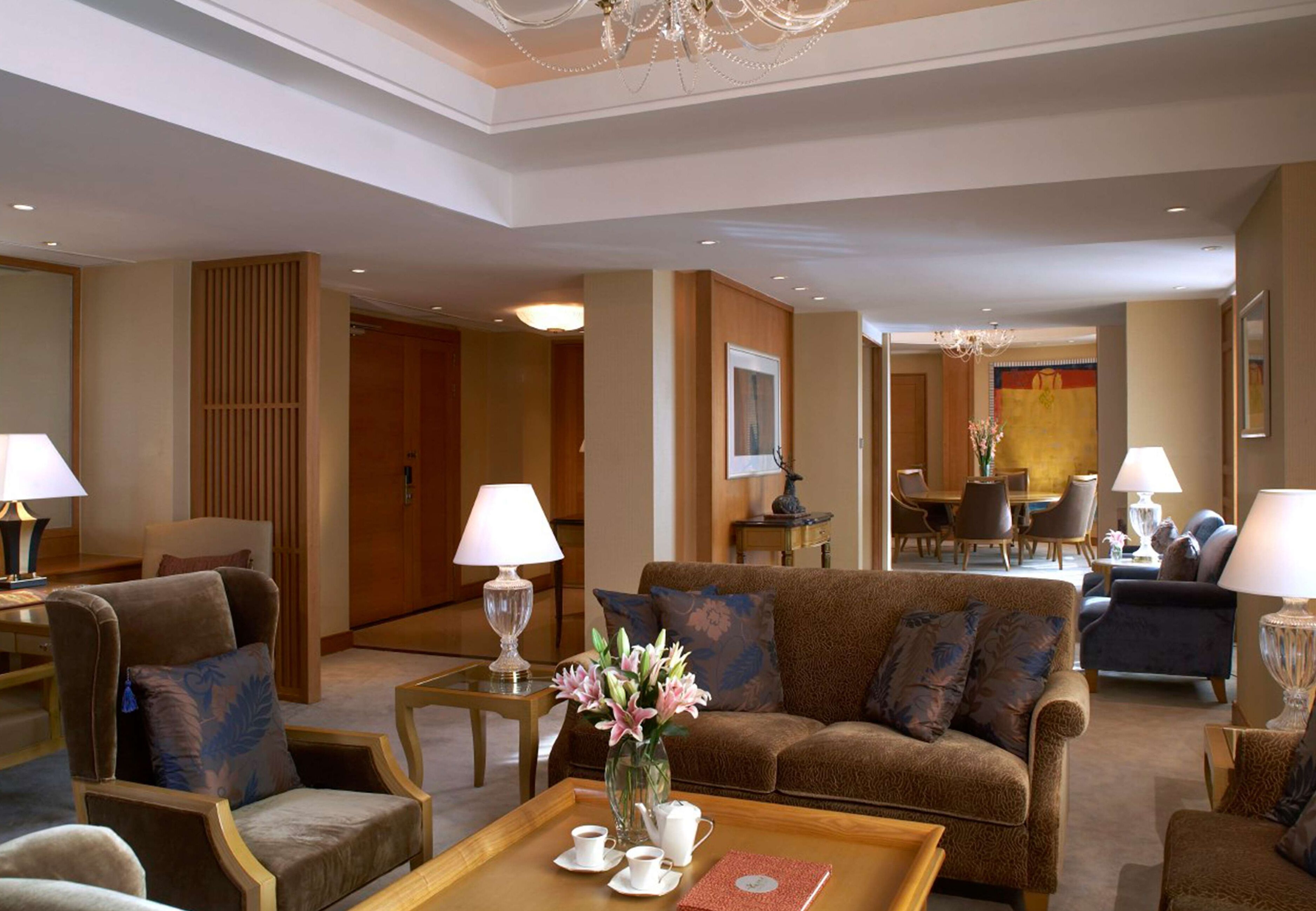 Un amplio vestíbulo de hotel con un sofá marrón, sillones, una mesa de centro con tazas y un jarrón de flores, además de una araña.