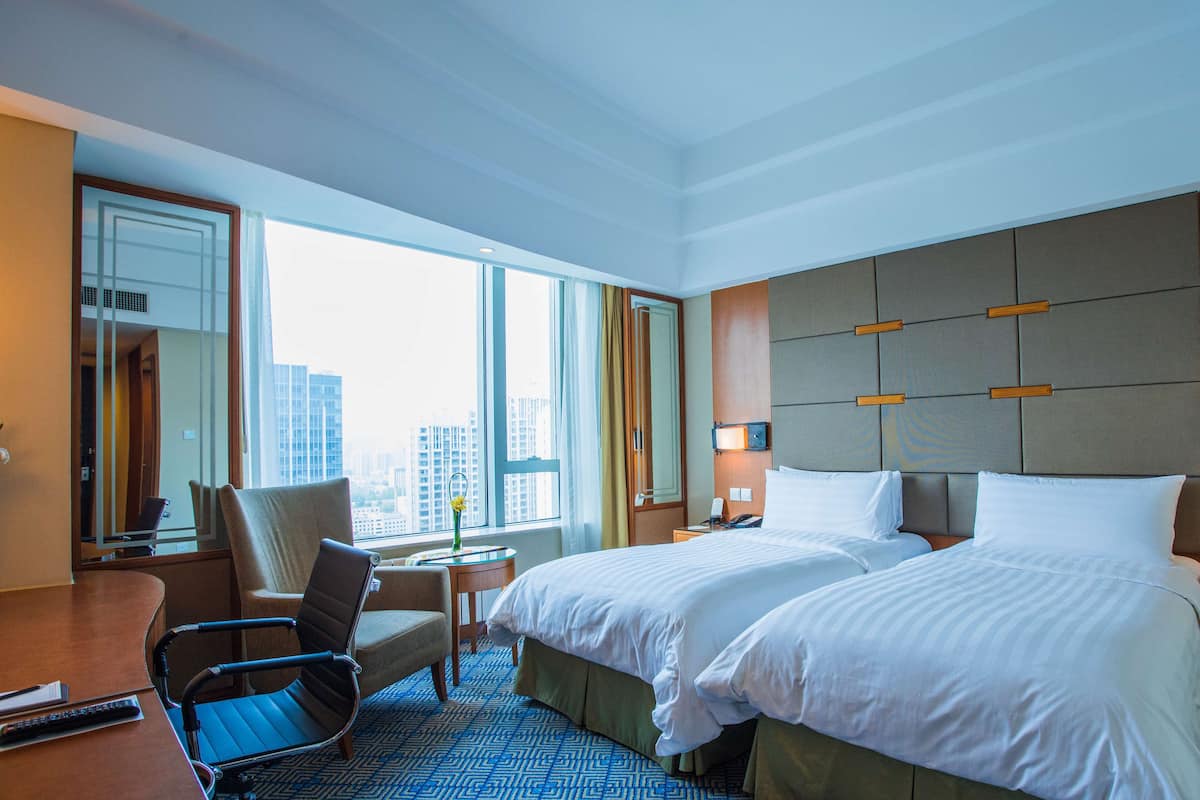 A hotel room with two beds, a desk, a chair, a large window with a city view, and a modern design.