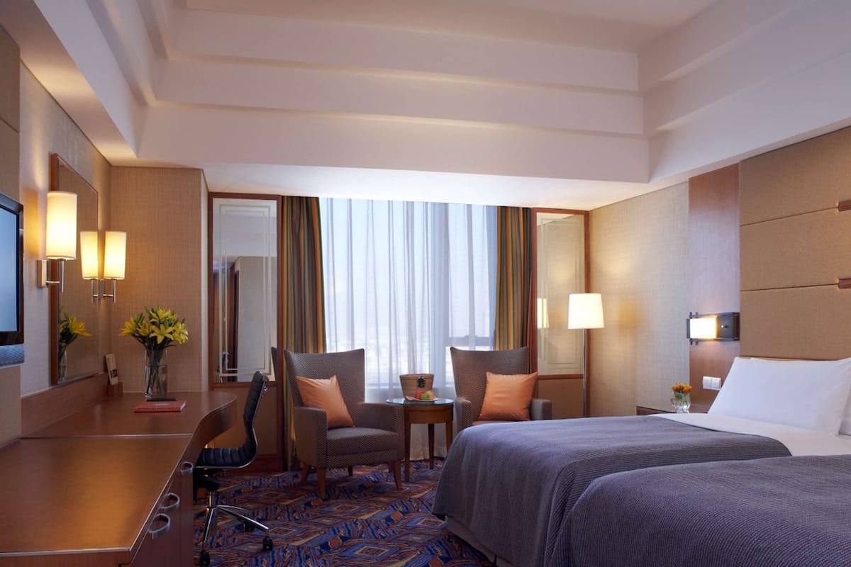 A hotel room with a large bed, two armchairs, a desk with a lamp, a TV, and a view of the outdoors through a window with curtains.