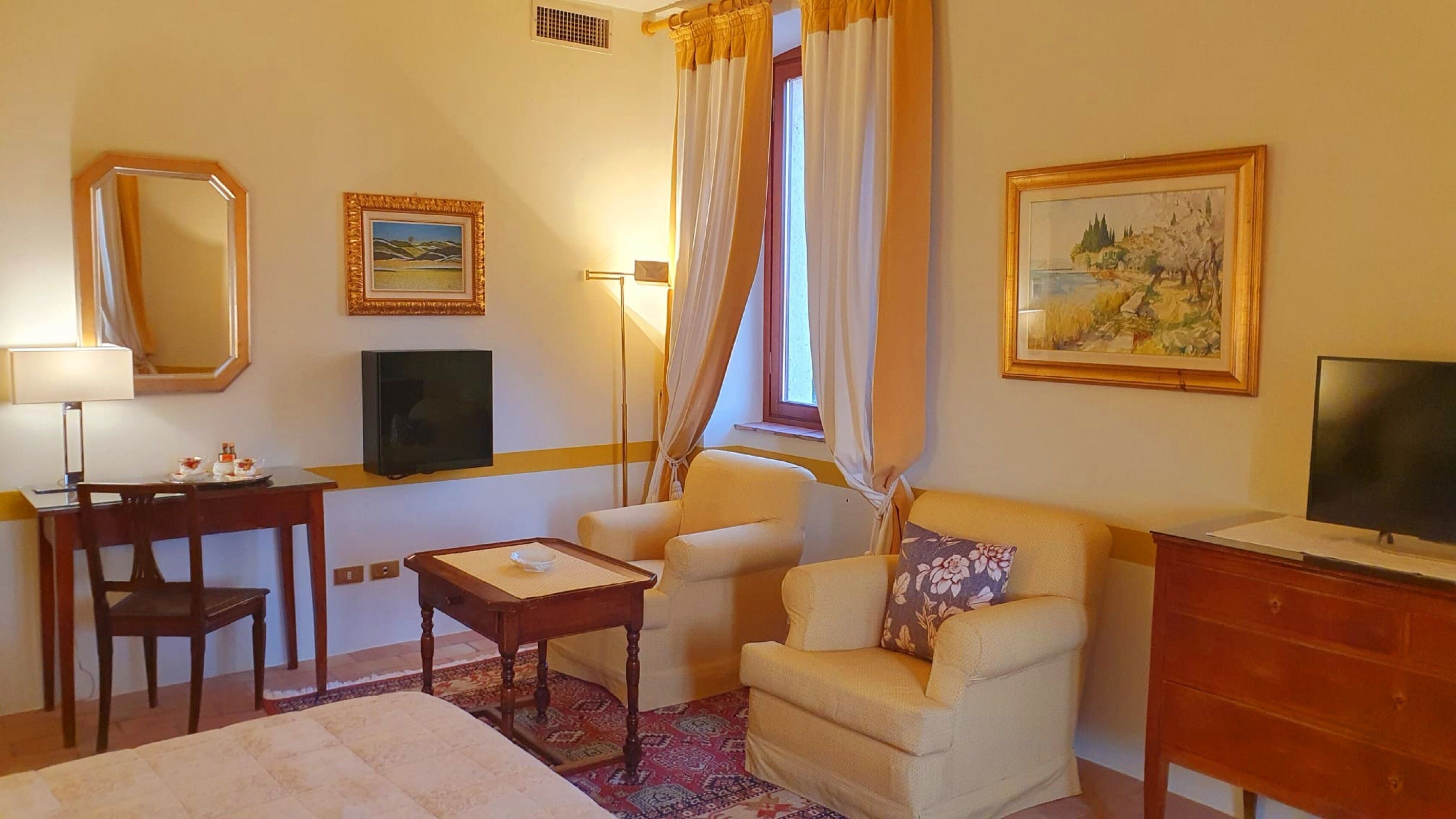 A hotel room with a bed, a desk with a lamp, a television, a chair, a sofa, and two paintings on the wall.