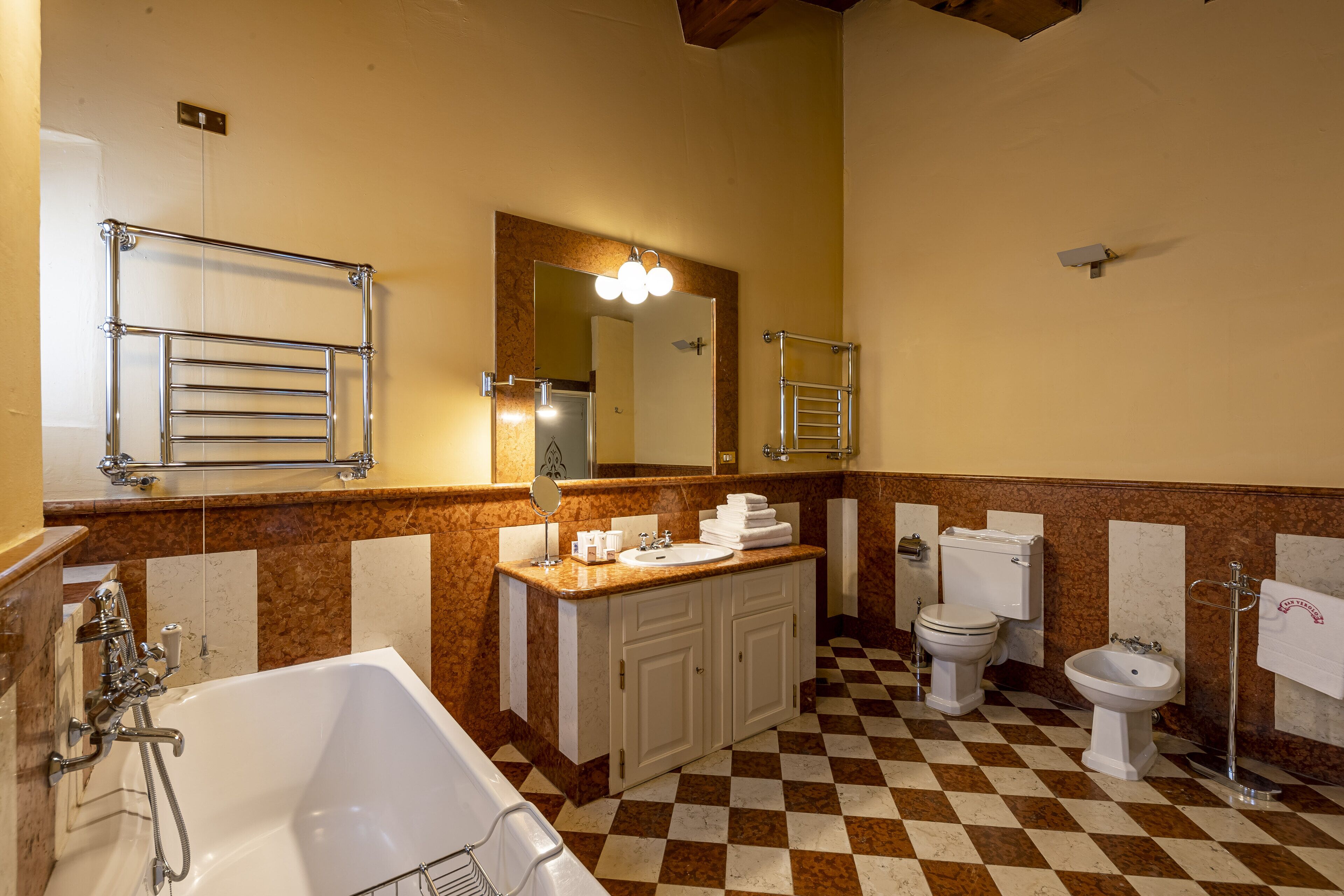 A bathroom with a checkered floor, a sink, a toilet, a bidet, a bathtub, and a mirror with lights.