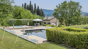 A swimming pool with lounge chairs and umbrellas, surrounded by well-manicured hedges and trees, with a view of distant hills. - Resort Locanda San Verolo Boutique Hotel (Costermano sul Garda)