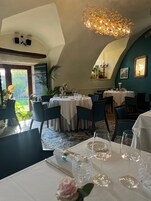 Un comedor con mesas dispuestas para dos, una araña y vistas al verde exterior.