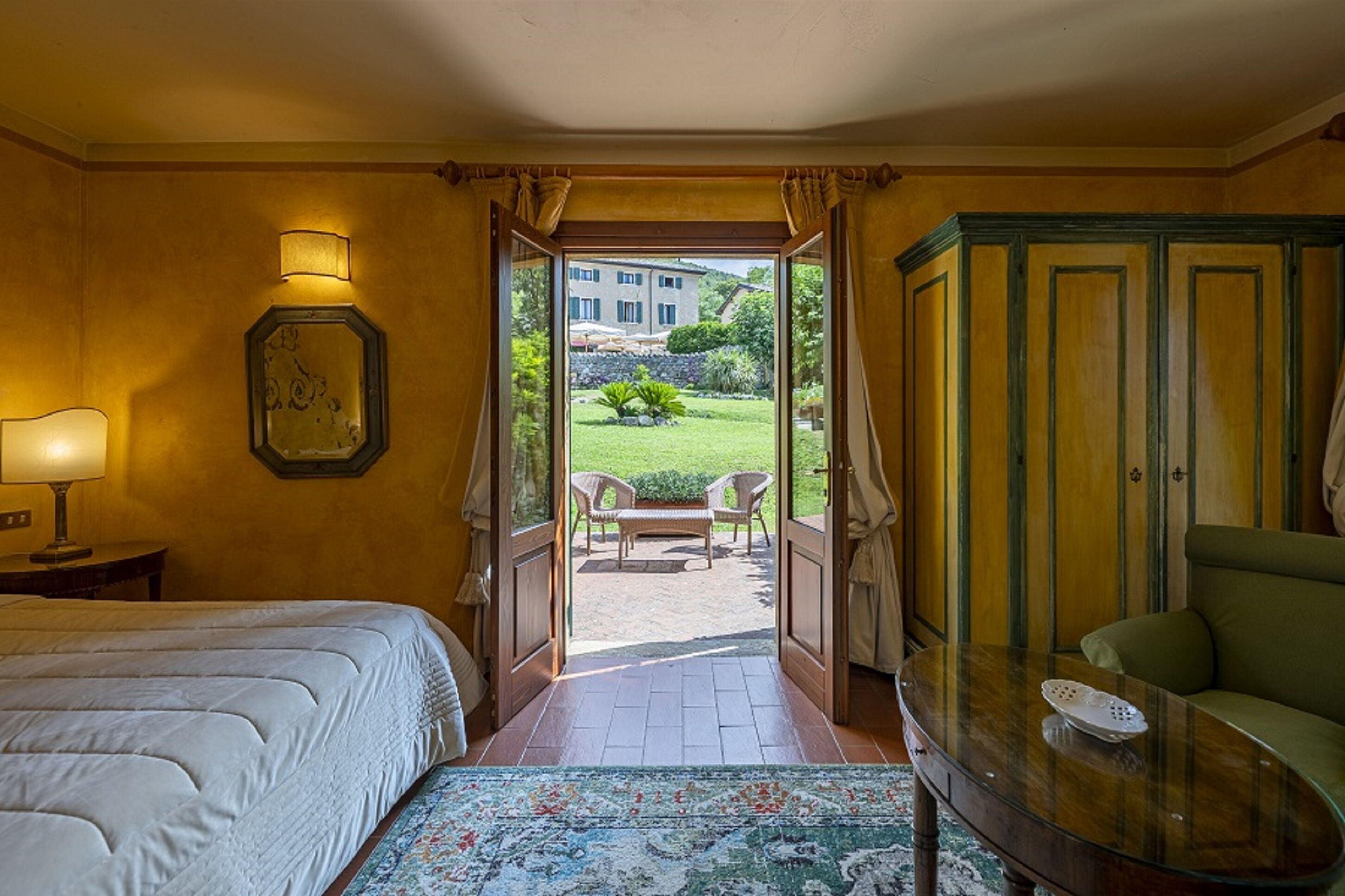 A hotel room with a bed, a nightstand, a lamp, a mirror, a green sofa, a glass table, and an open door leading to an outdoor seating area with chairs and a view of a building and greenery.