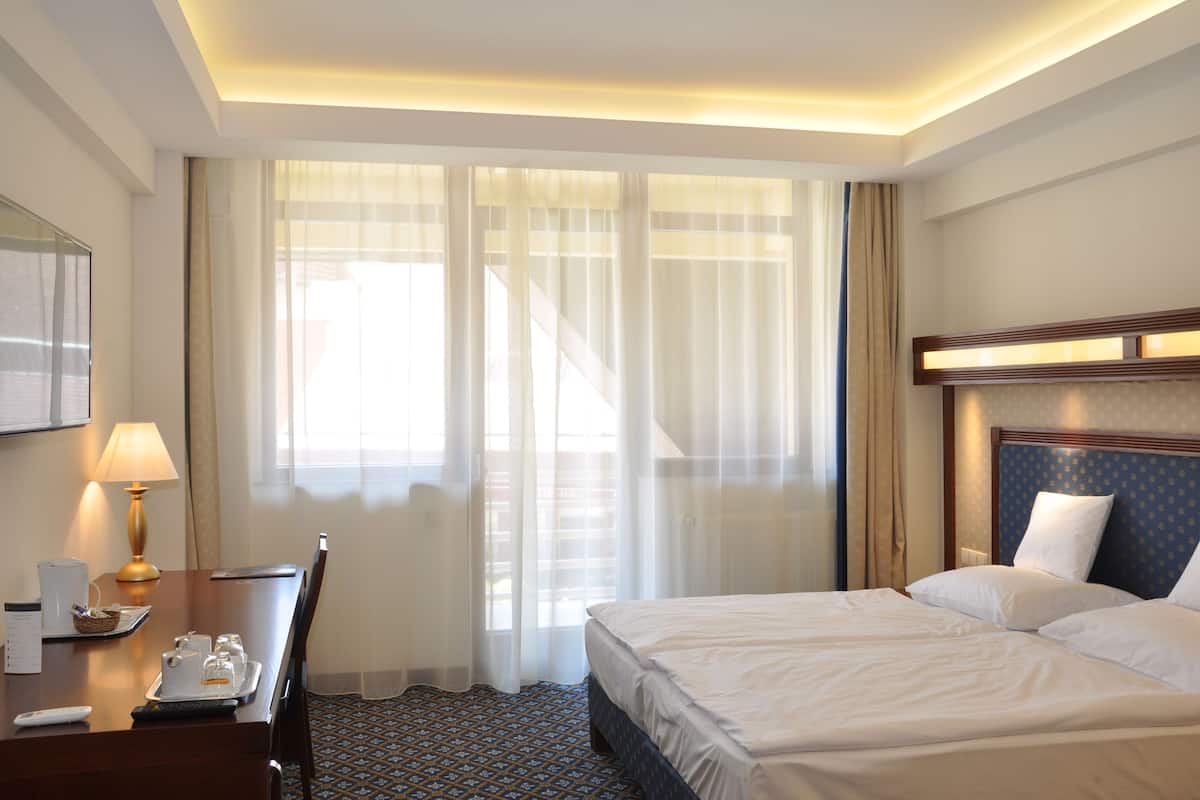 Ein Hotelzimmer mit Bett, Schreibtisch mit Lampe, Stuhl und Fenster mit durchsichtigen Vorhängen.