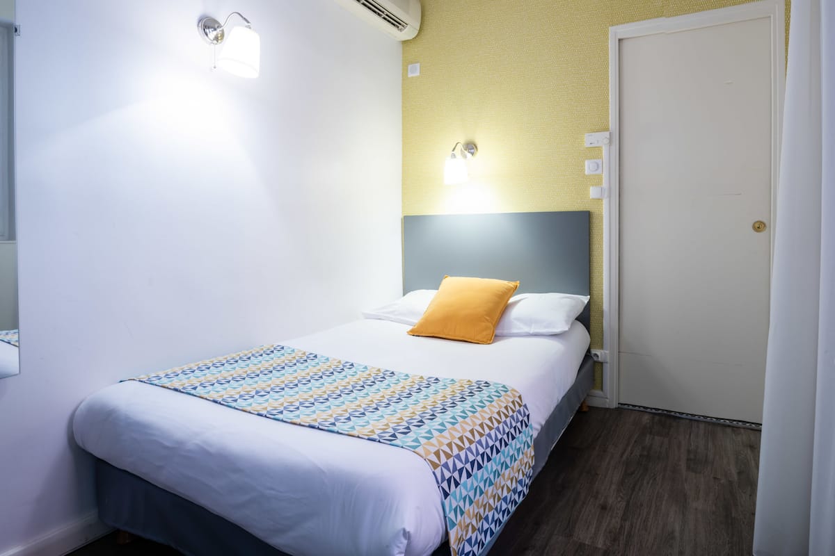 A neatly made bed with a patterned bedspread, a single yellow pillow, and a headboard against a textured wall.