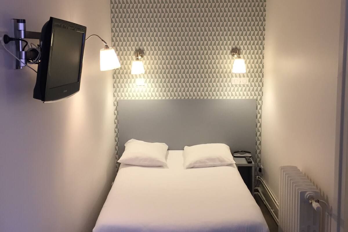 A small, neatly arranged bedroom with a bed, two bedside lamps, a mounted TV, and a radiator.