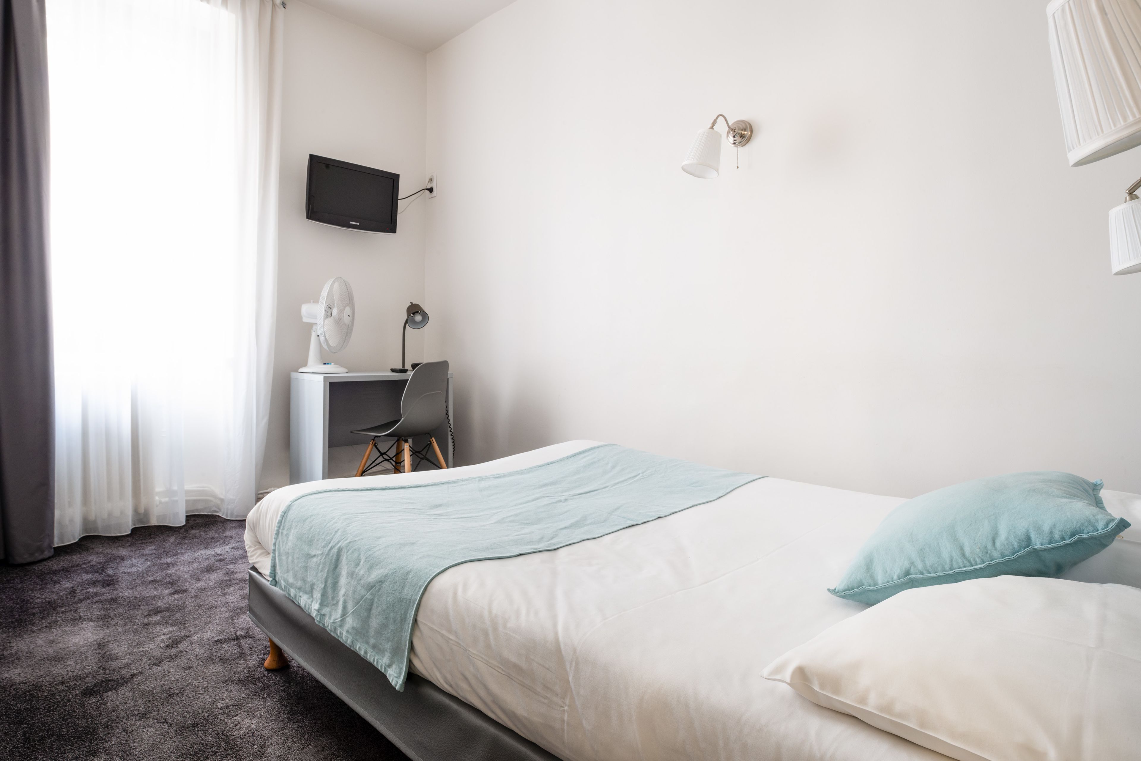 A bedroom with a bed, a desk, a chair, a TV, and a lamp.