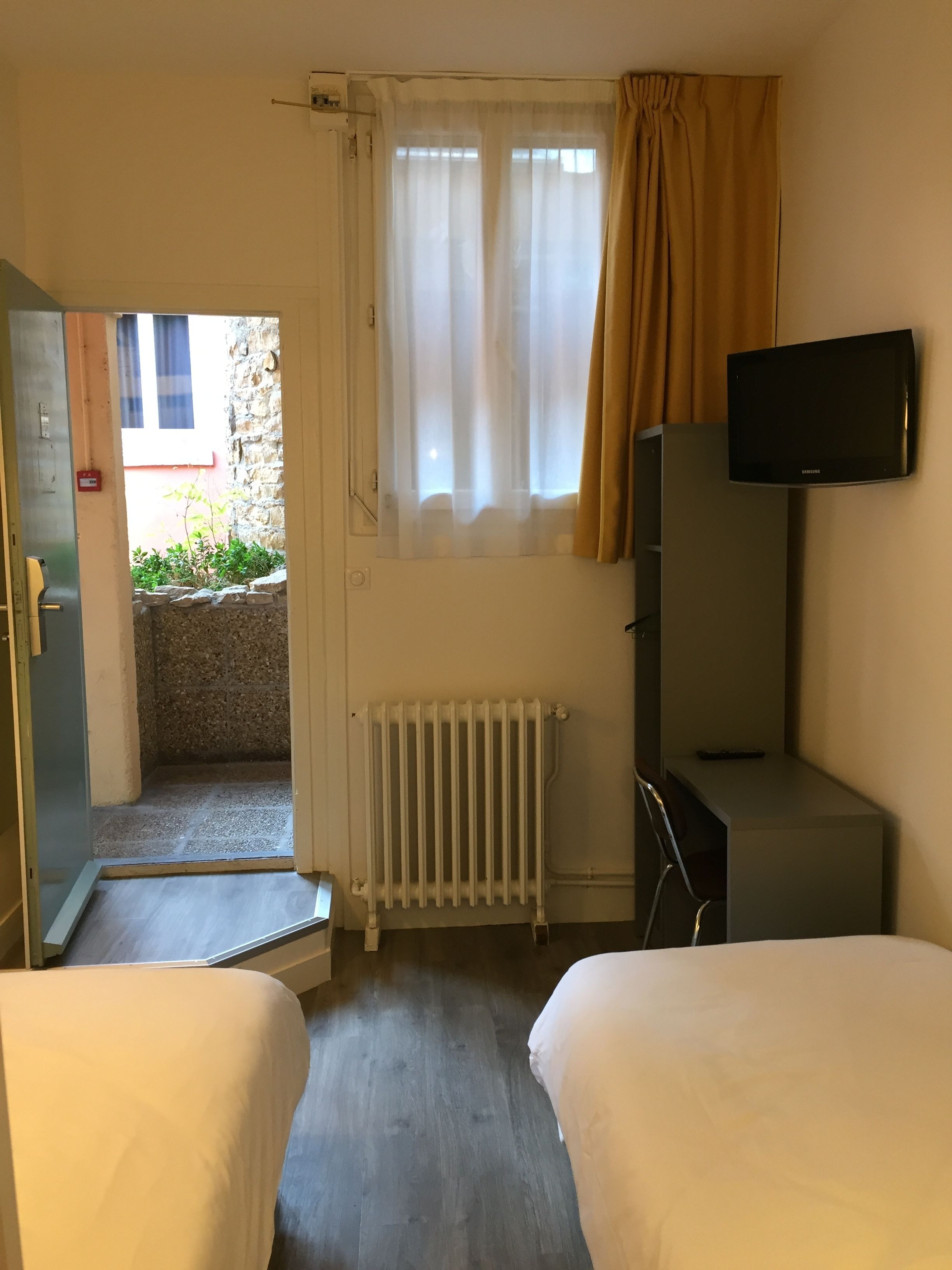 Camera d'albergo con due letti, televisione, finestra con tende, termosifone e vista su uno spazio esterno con piante.