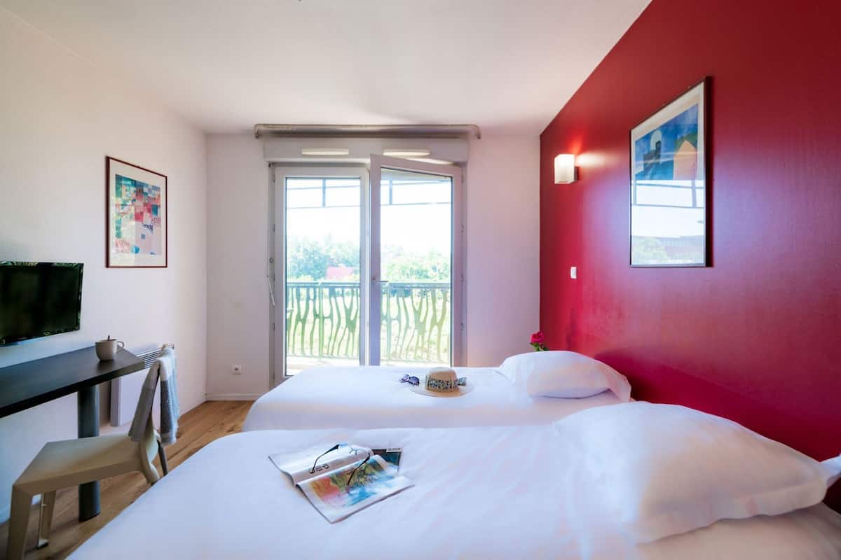 Habitación de hotel con dos camas, televisión, escritorio y balcón con vistas.