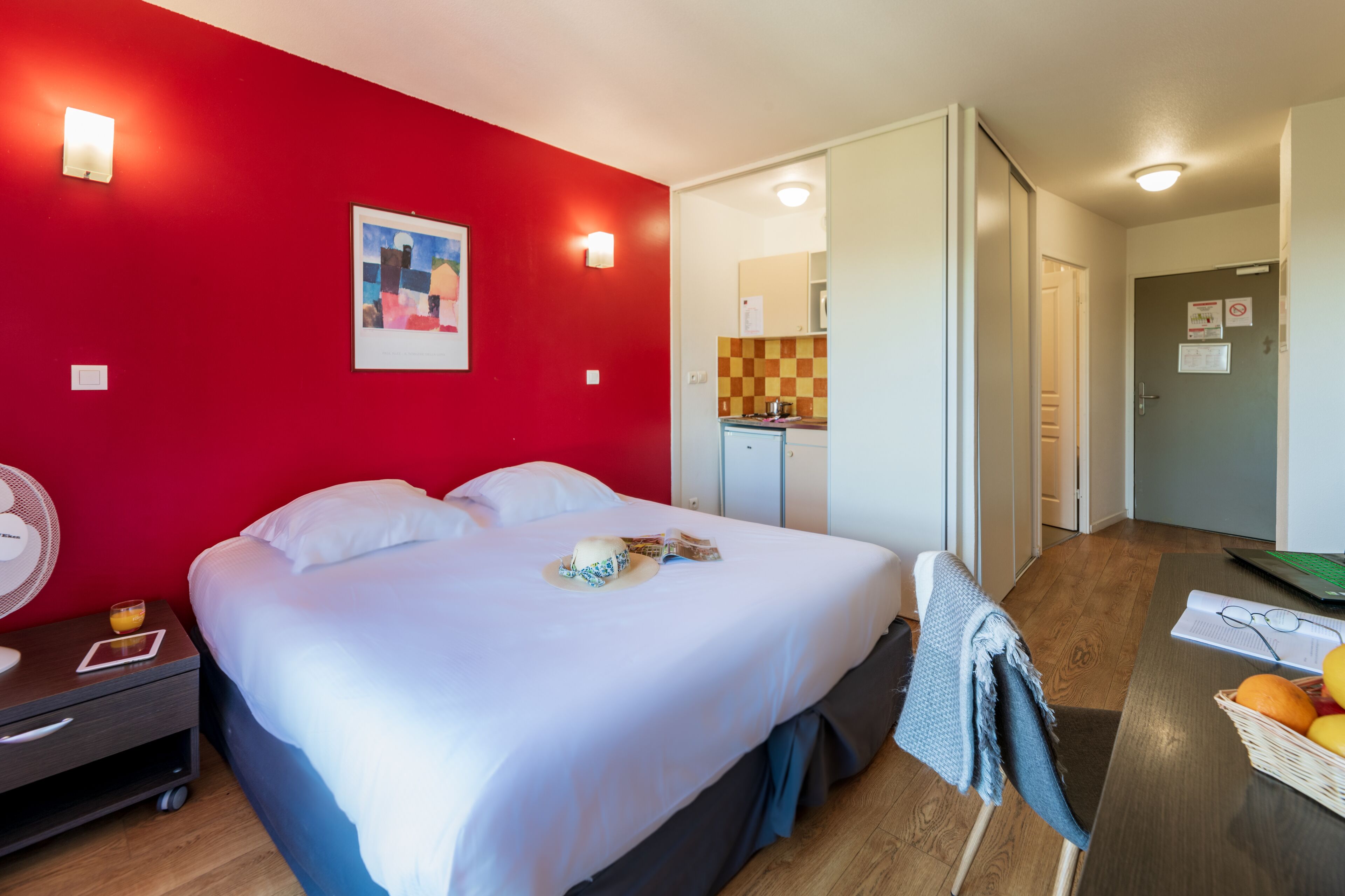 Une chambre d’hôtel comprenant un lit, une table de chevet, un ventilateur, une table à manger avec des fruits et un coin cuisine.