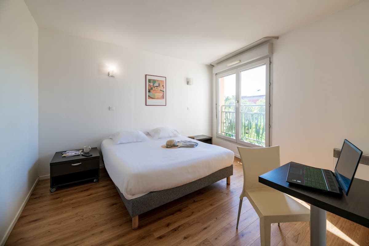 Un dormitorio con cama, escritorio con ordenador portátil, mesita de noche, silla y ventana con vistas.