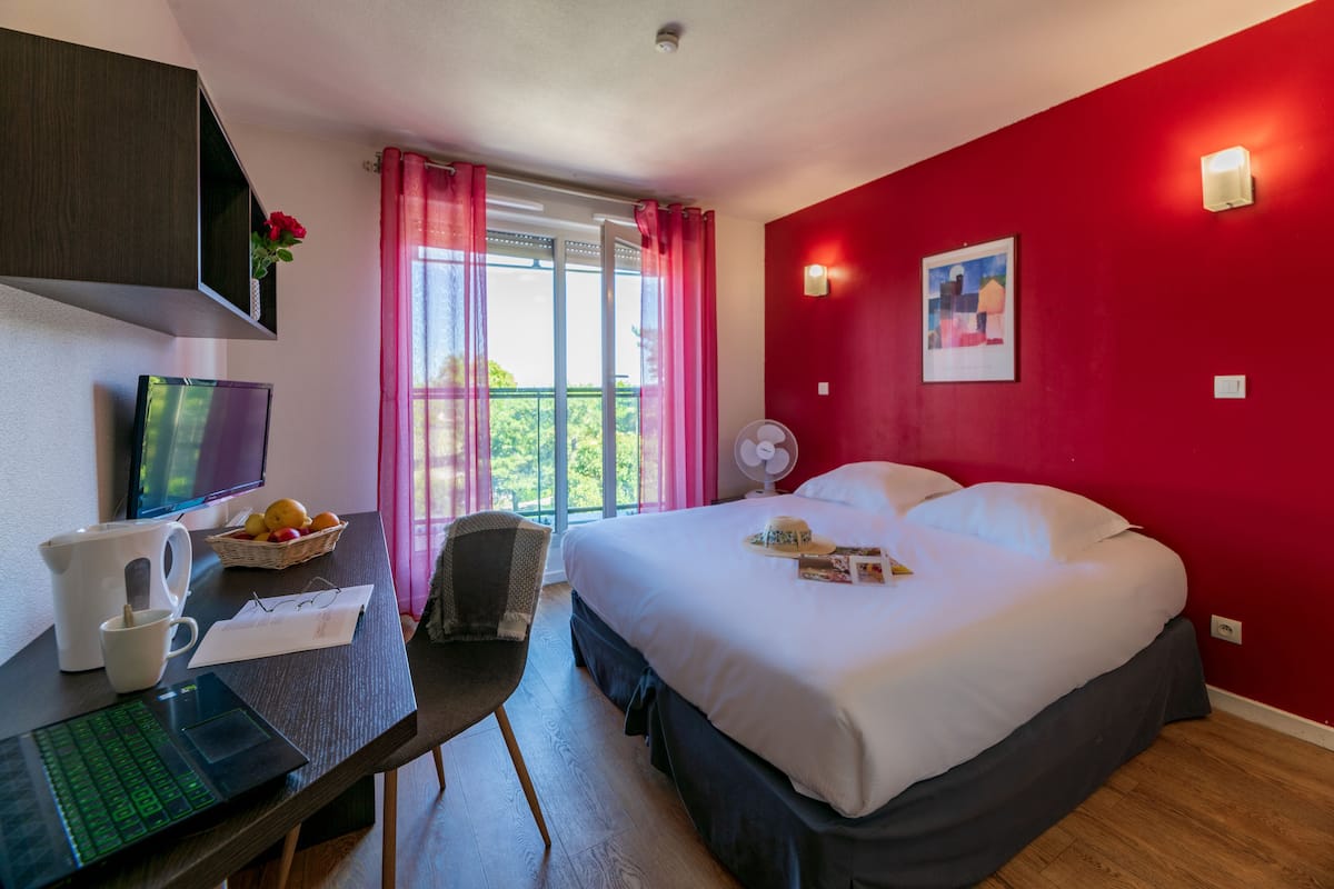 Habitación de hotel con cama, escritorio con computadora, silla, ventana con cortinas y ventilador en la pared.