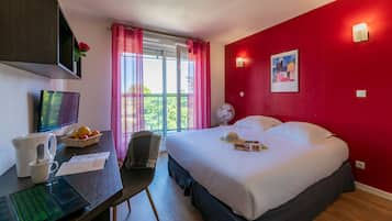 A hotel room with a bed, a desk with a computer, a chair, a window with curtains, and a wall-mounted fan.