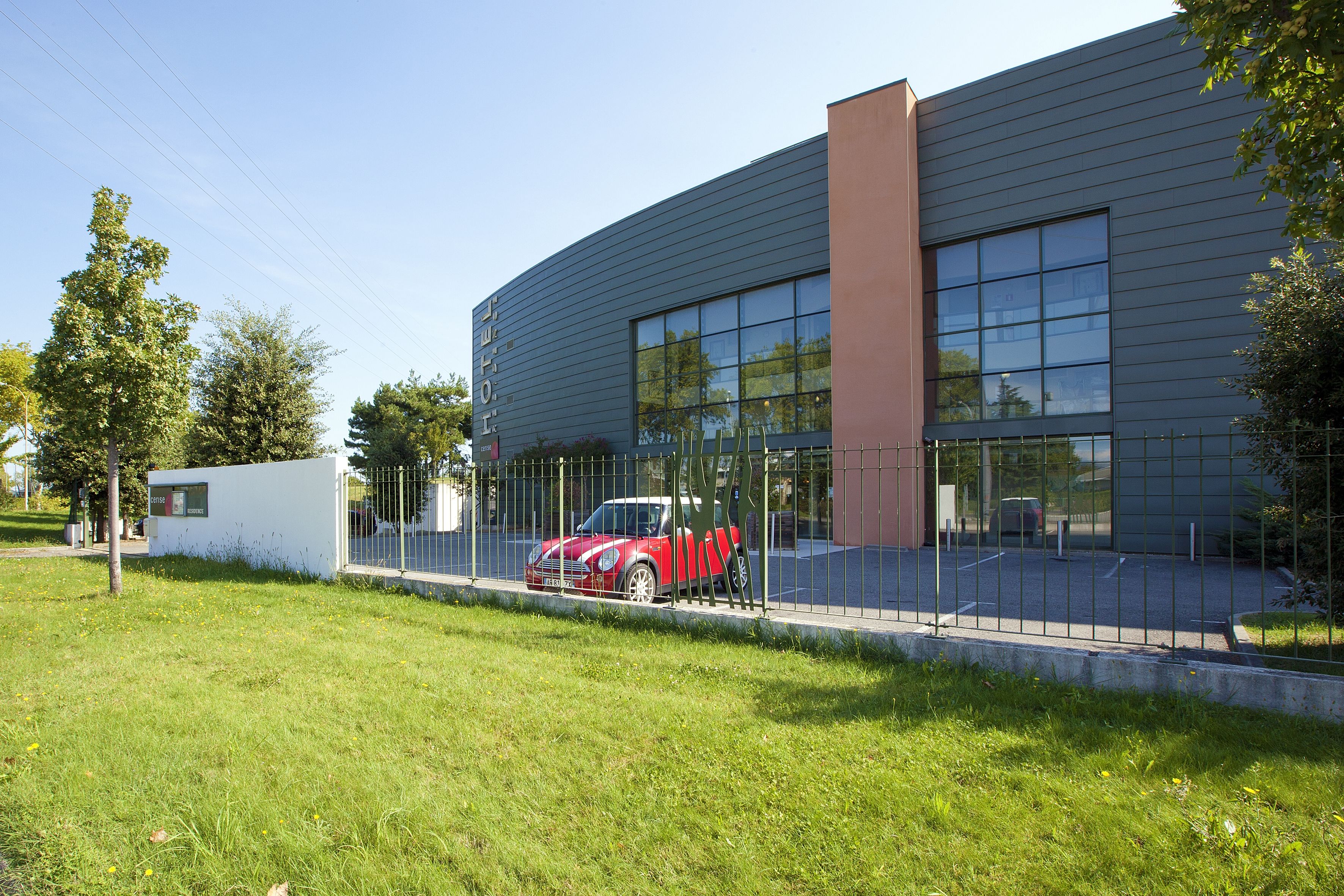 Un bâtiment moderne doté d’une grande façade en verre, une voiture rouge garée devant, et un espace verdoyant bien entretenu.