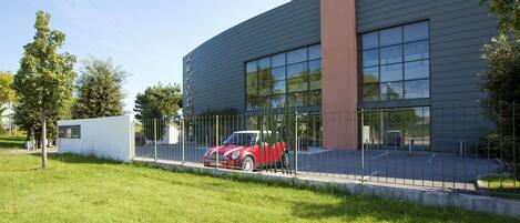 Un bâtiment moderne doté d’une grande façade en verre, une voiture rouge garée devant, et un espace verdoyant bien entretenu.