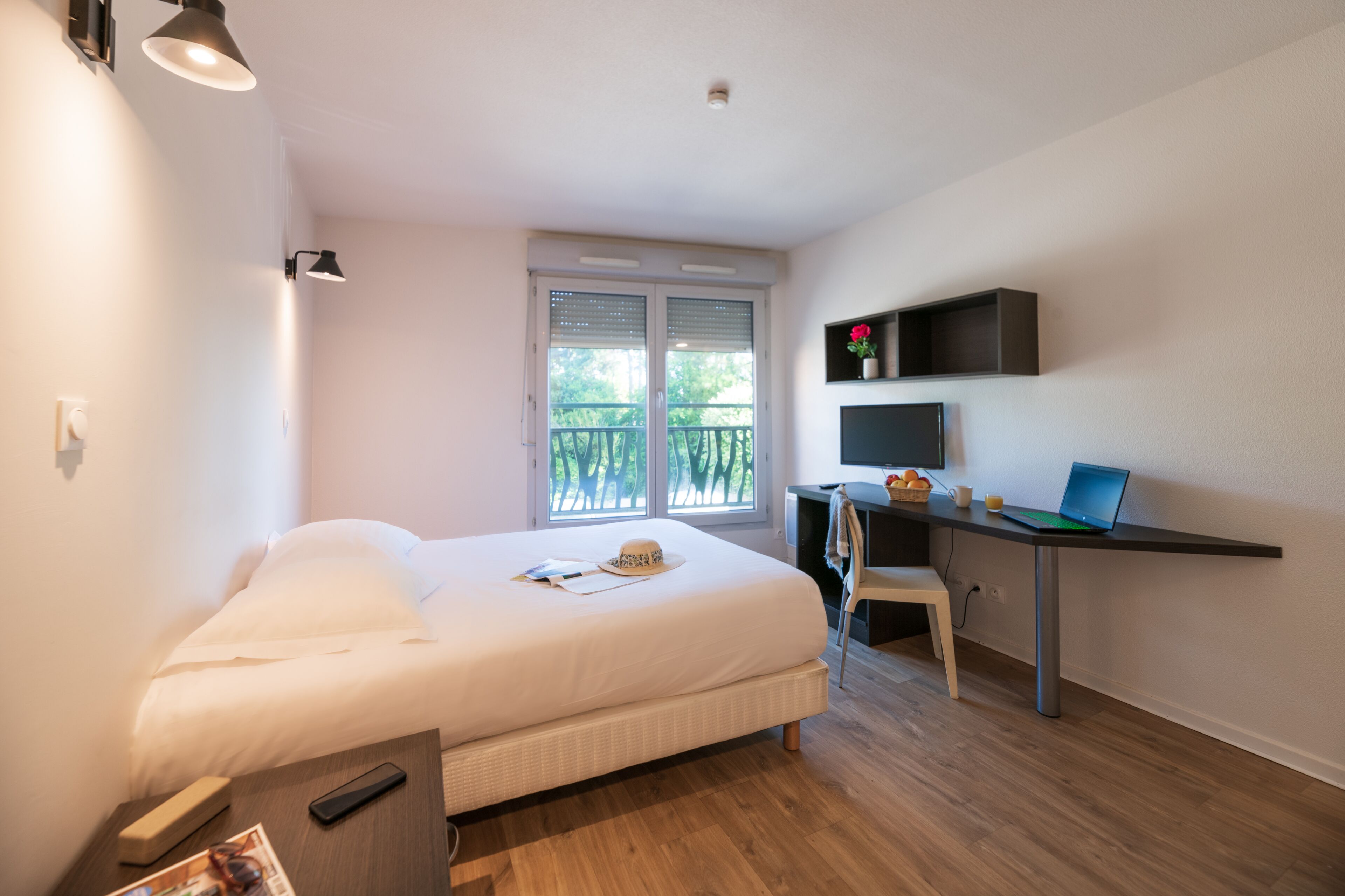 A bedroom with a bed, a desk with a laptop, a chair, a TV, and a window with blinds.