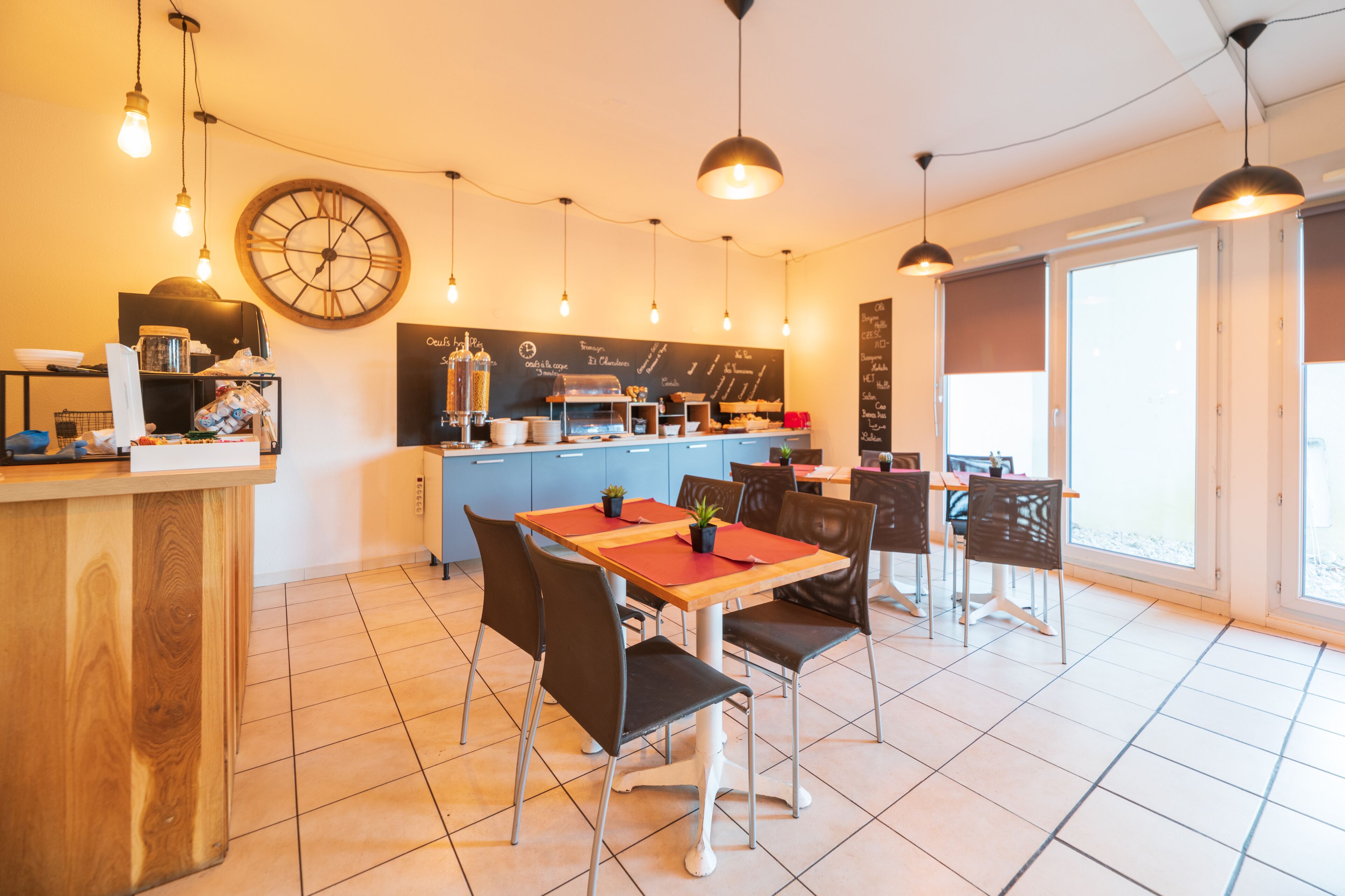 A modern cafe with a wooden counter, blackboard menu, and a large clock. There are tables and chairs for dining, and the space is well-lit with pendant lights.