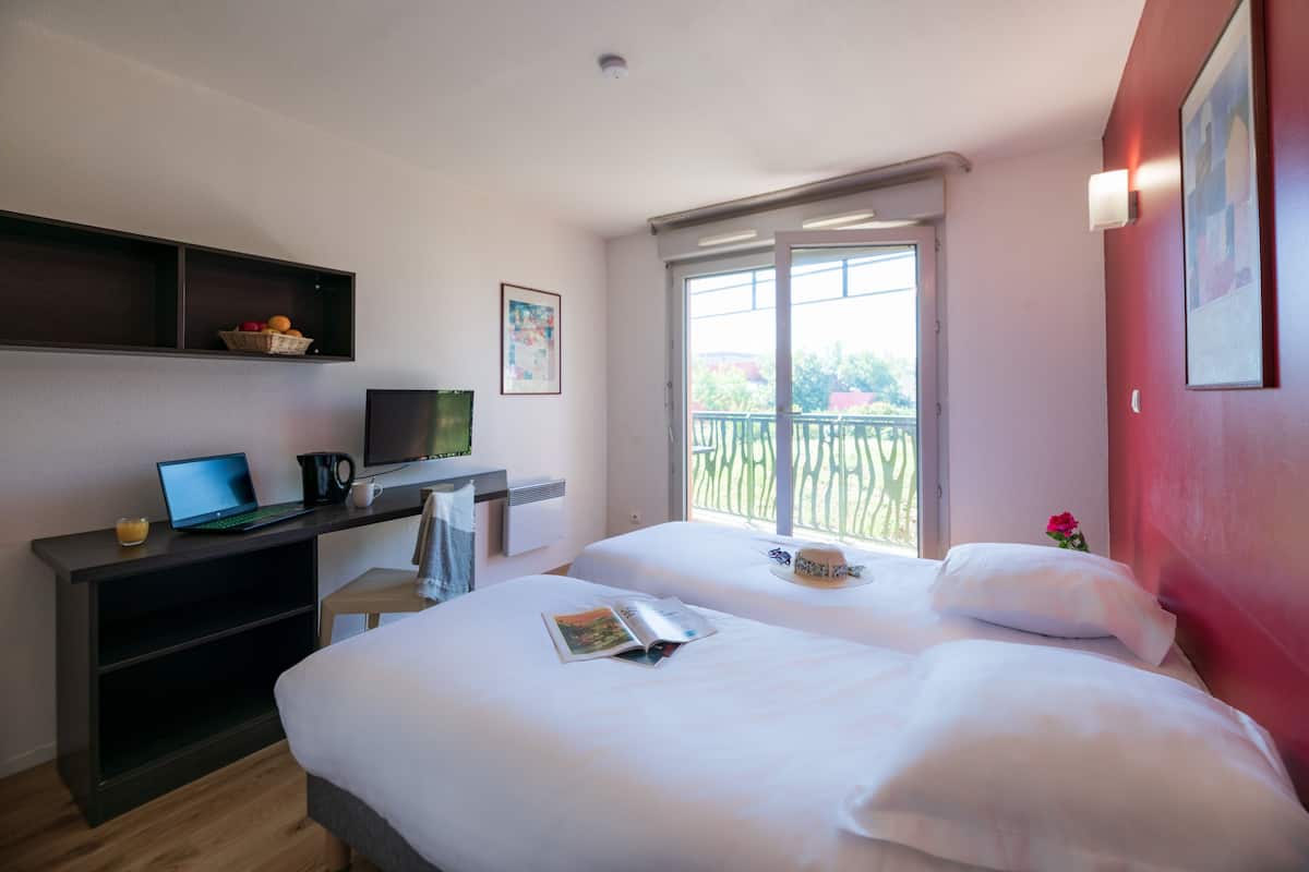 Habitación de hotel con dos camas, un escritorio con una laptop y una cafetera, una ventana con vista a follaje, y una pared de acento roja.