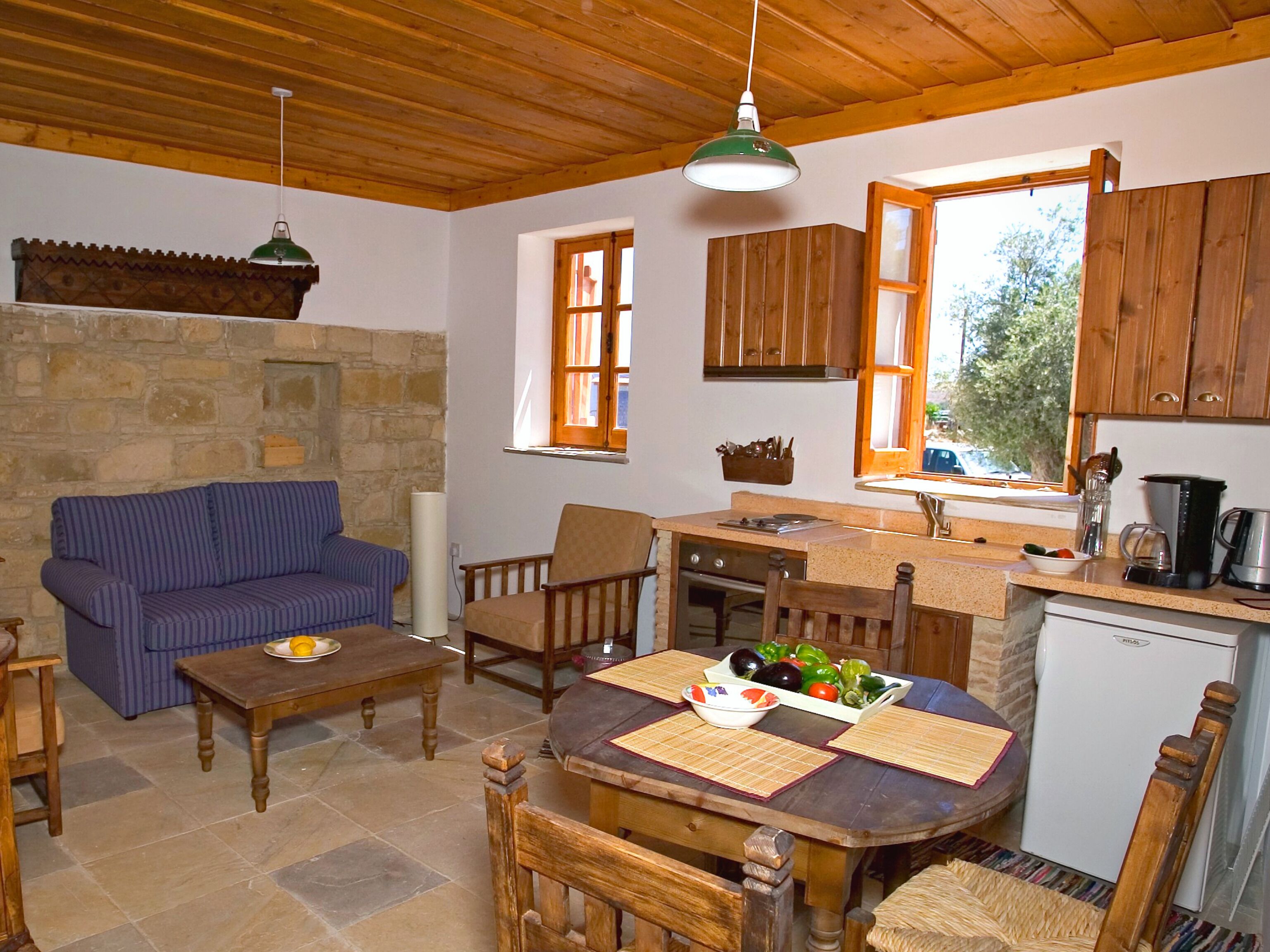 A cozy room with a stone wall, wooden furniture, a dining table with placemats, a sofa, and a kitchen area with wooden cabinets and a window.