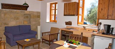 A cozy room with a stone wall, wooden furniture, a dining table with placemats, a sofa, and a kitchen area with wooden cabinets and a window.