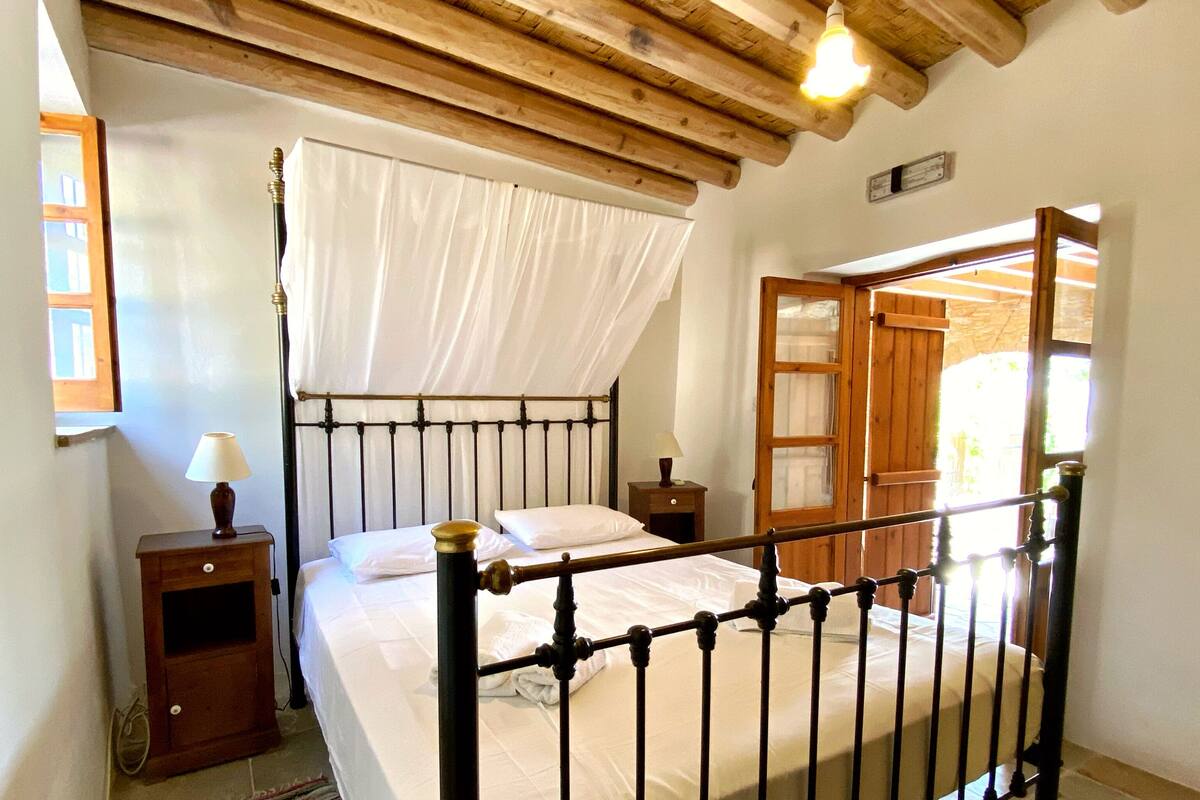 A bedroom with a wooden ceiling, a bed with a metal frame, a nightstand with a lamp, and a window with curtains.