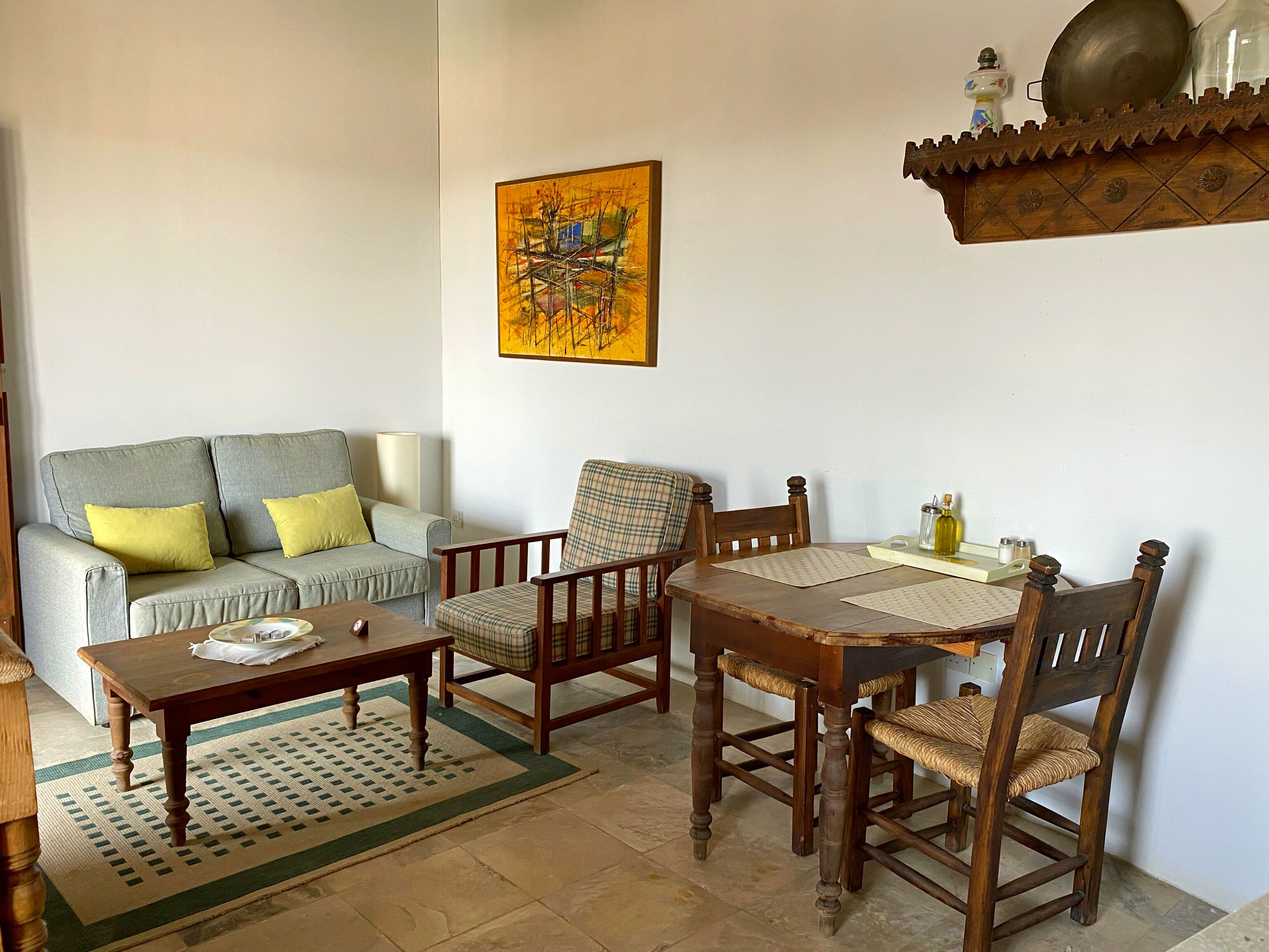 A living room with a grey sofa, two armchairs, a wooden coffee table, and a dining table with chairs. A painting hangs on the wall, and a shelf with decorative items is above the dining table.