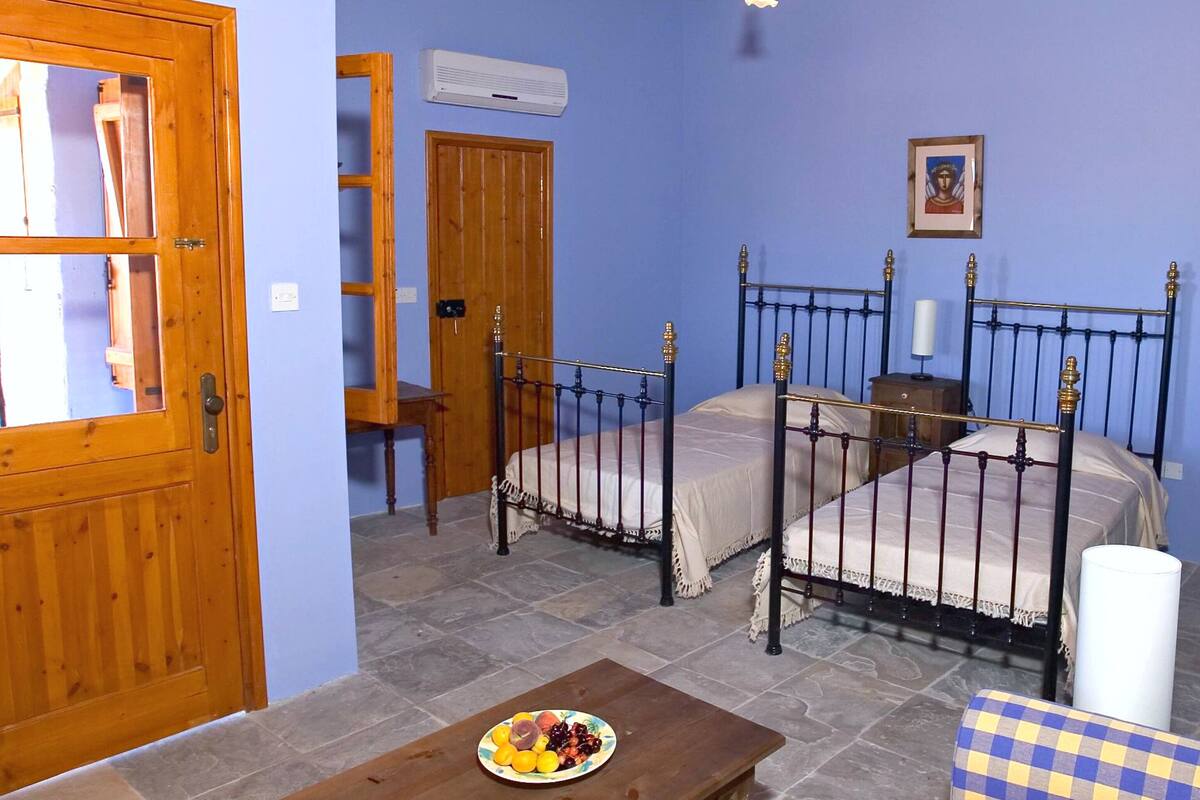 A bedroom with two beds, a wooden door, an air conditioning unit, a bedside table, and a fruit plate on the floor.