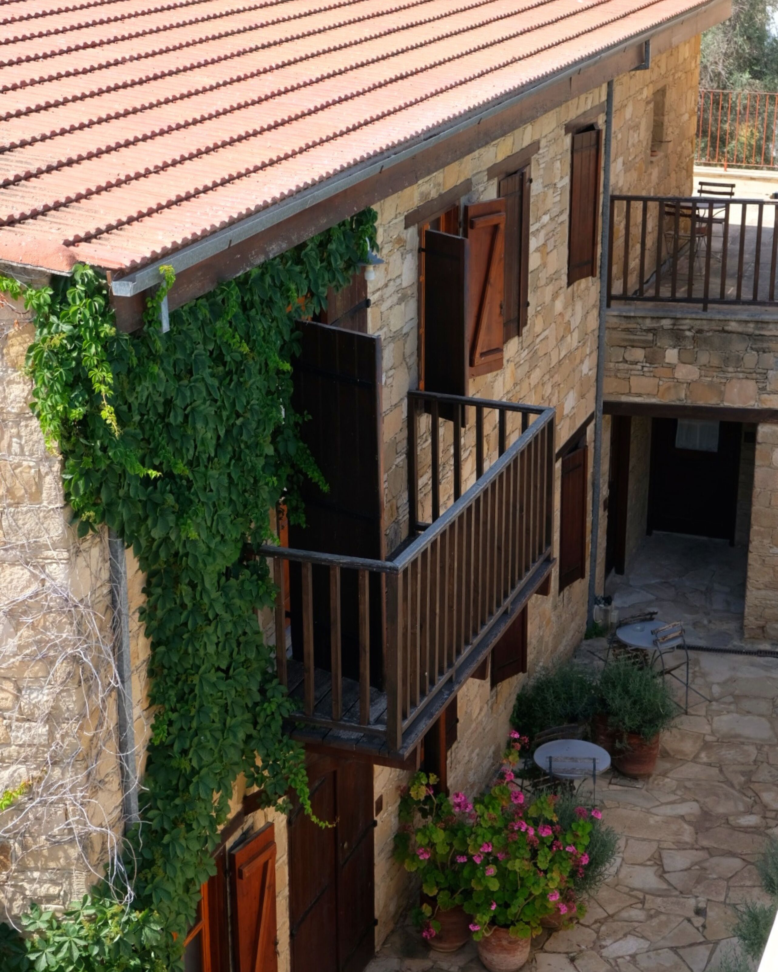 A stone building with a red-tiled roof, wooden shutters, and a balcony with a metal railing. There are potted plants and flowers on the ground.