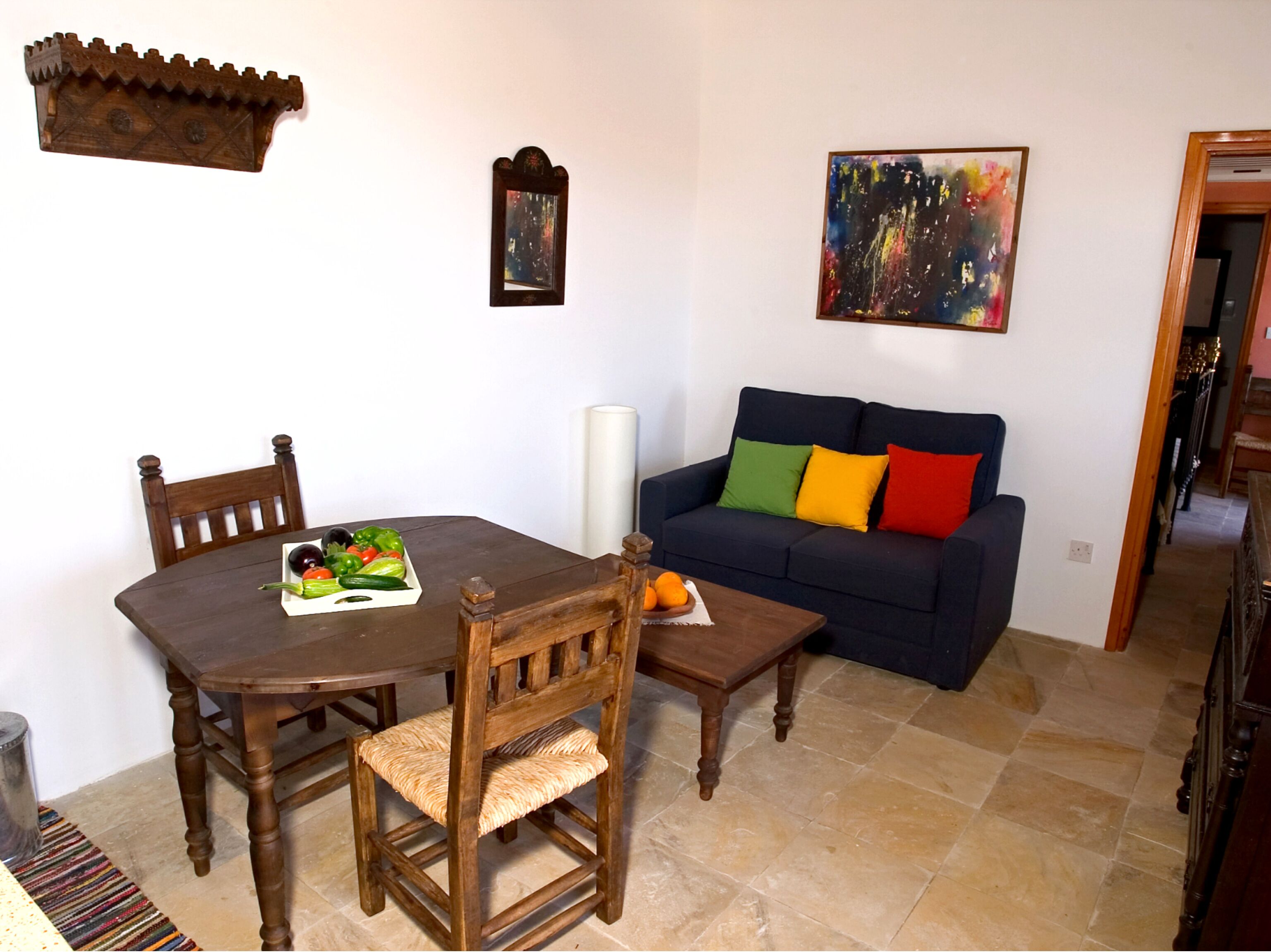 A dining area with a wooden table and chairs, a blue sofa with colorful cushions, and a painting on the wall.