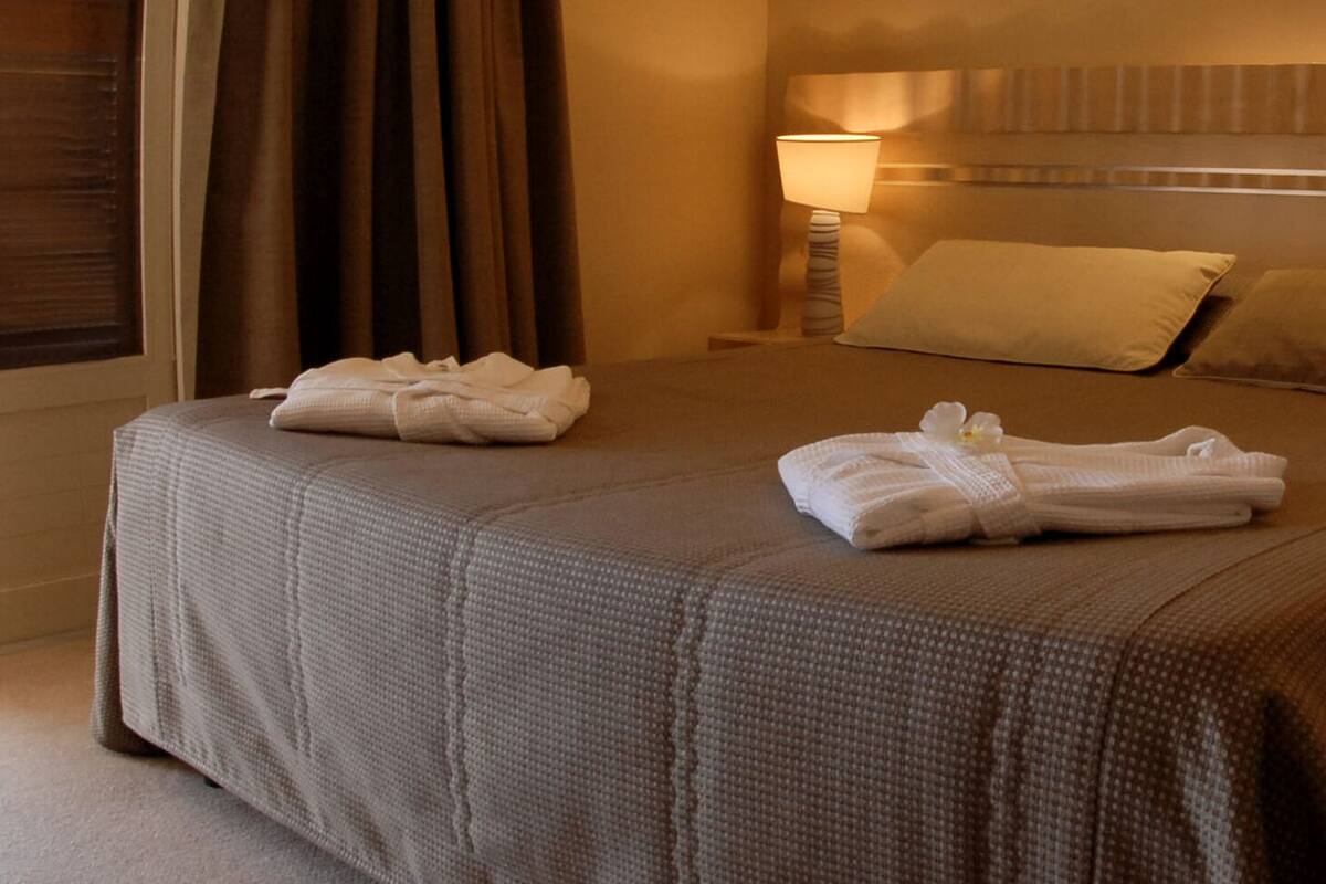 A neatly made bed with two towels folded and placed on it, a pillow, and a bedside lamp on a wooden nightstand.