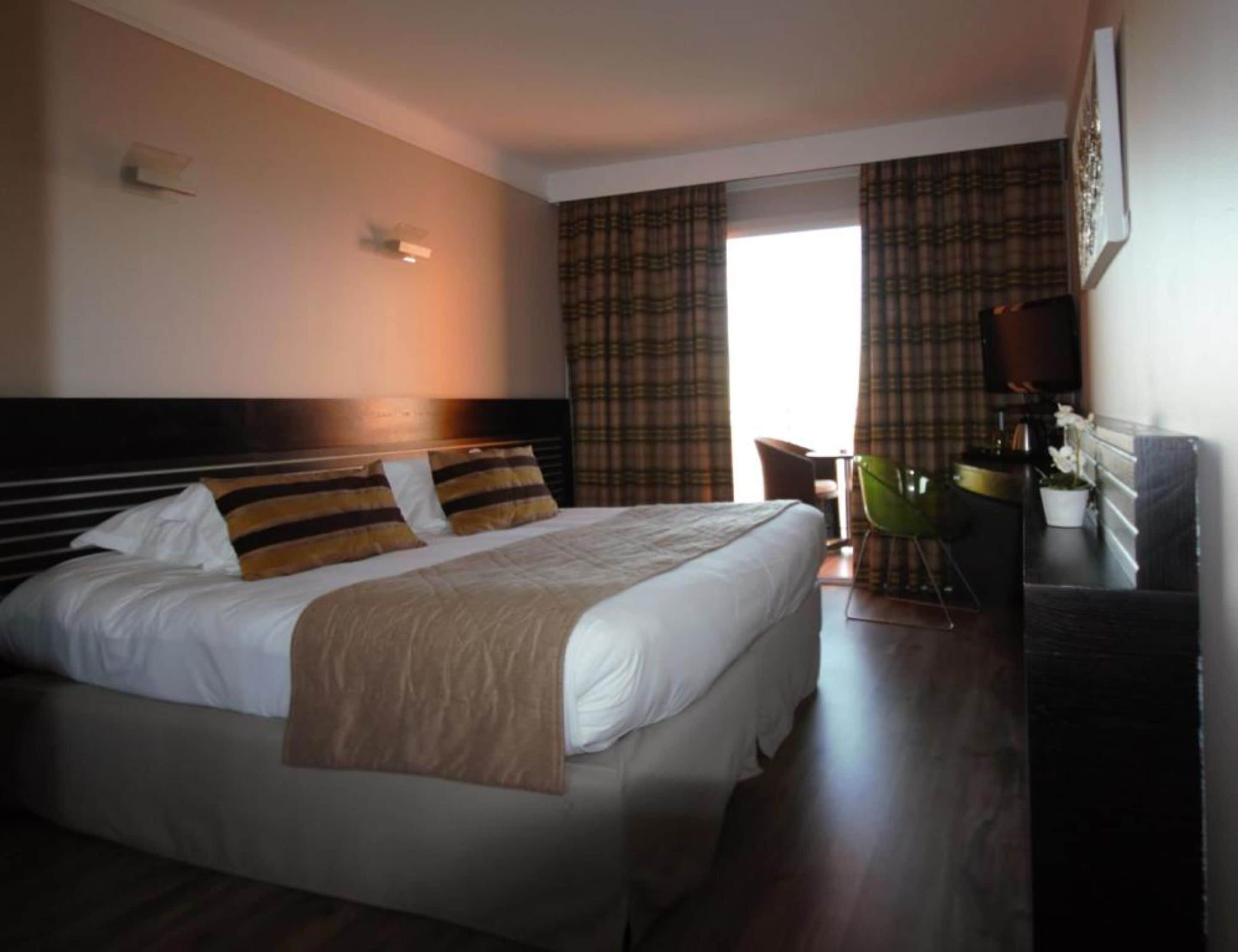 Une chambre d’hôtel avec un grand lit, un bureau avec une chaise, une télévision et une fenêtre avec des rideaux.