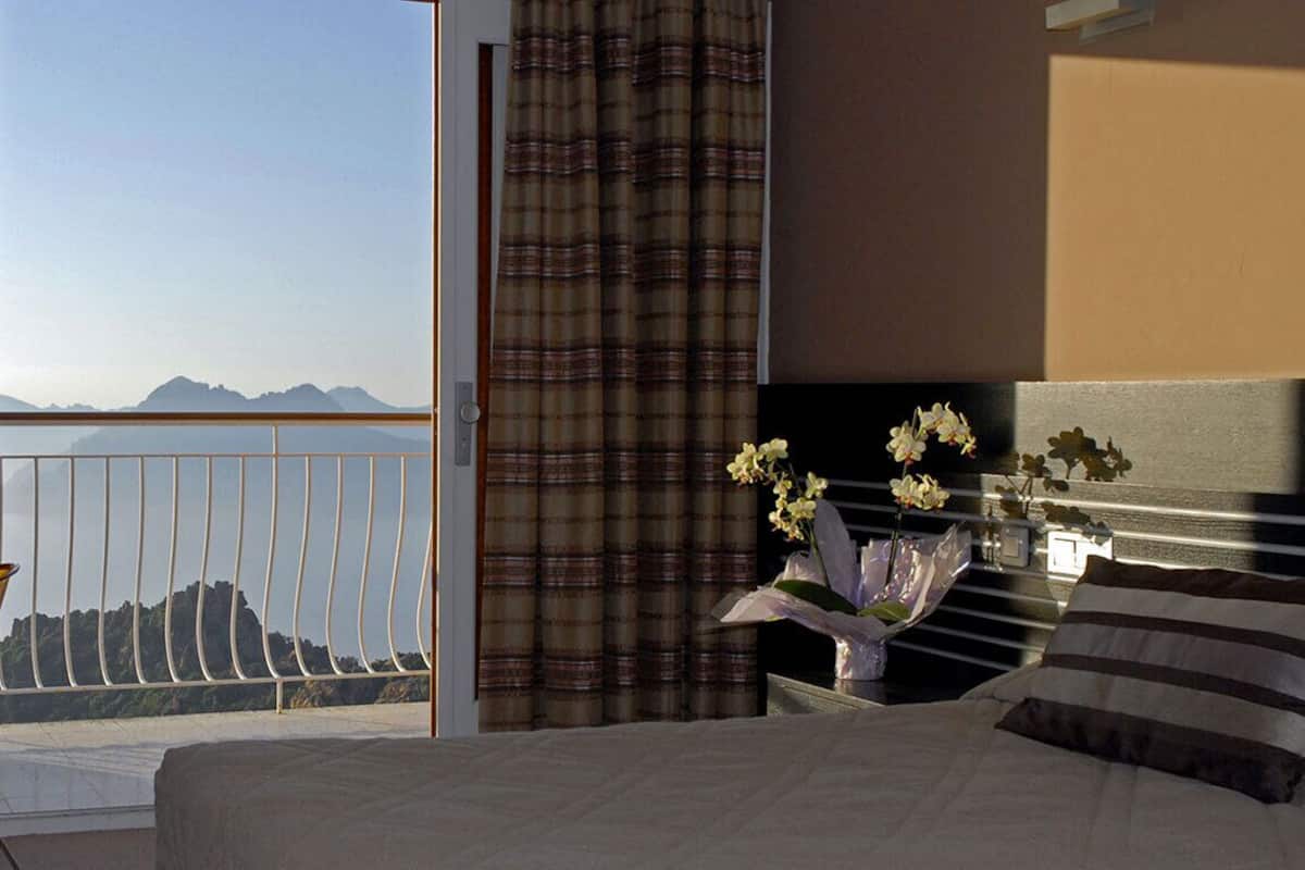 A hotel room with a bed, a nightstand with a vase of flowers, a window with curtains, and a view of mountains.