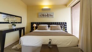Habitación de hotel con una cama grande, una mesita de noche con lámpara, un cuadro sobre la cama y una ventana con cortinas.