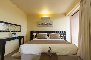 Habitación de hotel con una cama grande, una mesita de noche con lámpara, un cuadro sobre la cama y una ventana con cortinas.