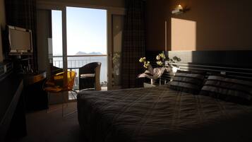 A hotel room with a large bed, a desk with a TV, a chair, and a view of the sea.