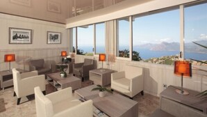 Un moderno vestíbulo de hotel con un amplio ventanal que ofrece una vista panorámica del mar y las montañas. Hay varios sofás y sillones dispuestos para los huéspedes.