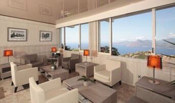 Un moderno vestíbulo de hotel con un amplio ventanal que ofrece una vista panorámica del mar y las montañas. Hay varios sofás y sillones dispuestos para los huéspedes.