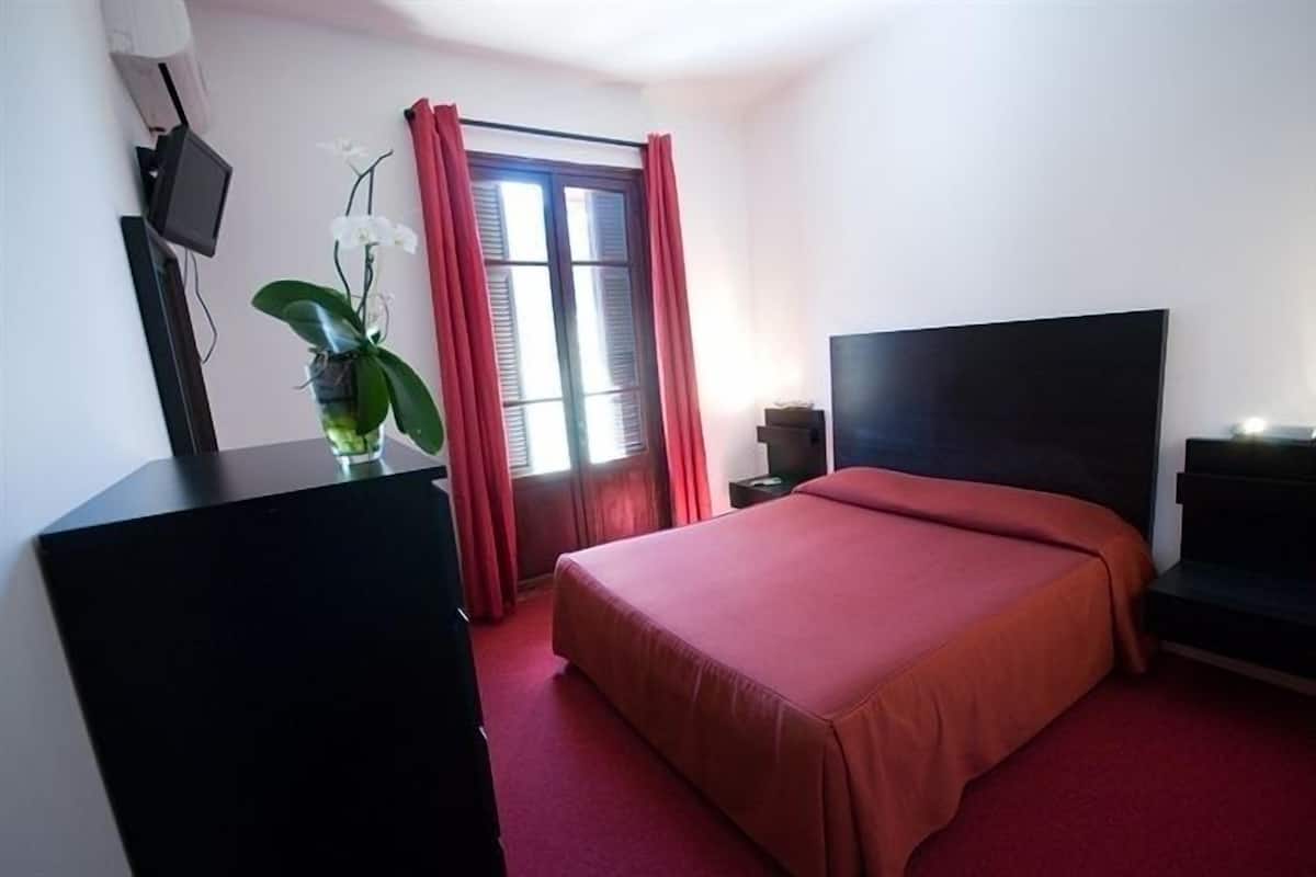 A hotel room with a bed, a TV, a desk, and a window with curtains.
