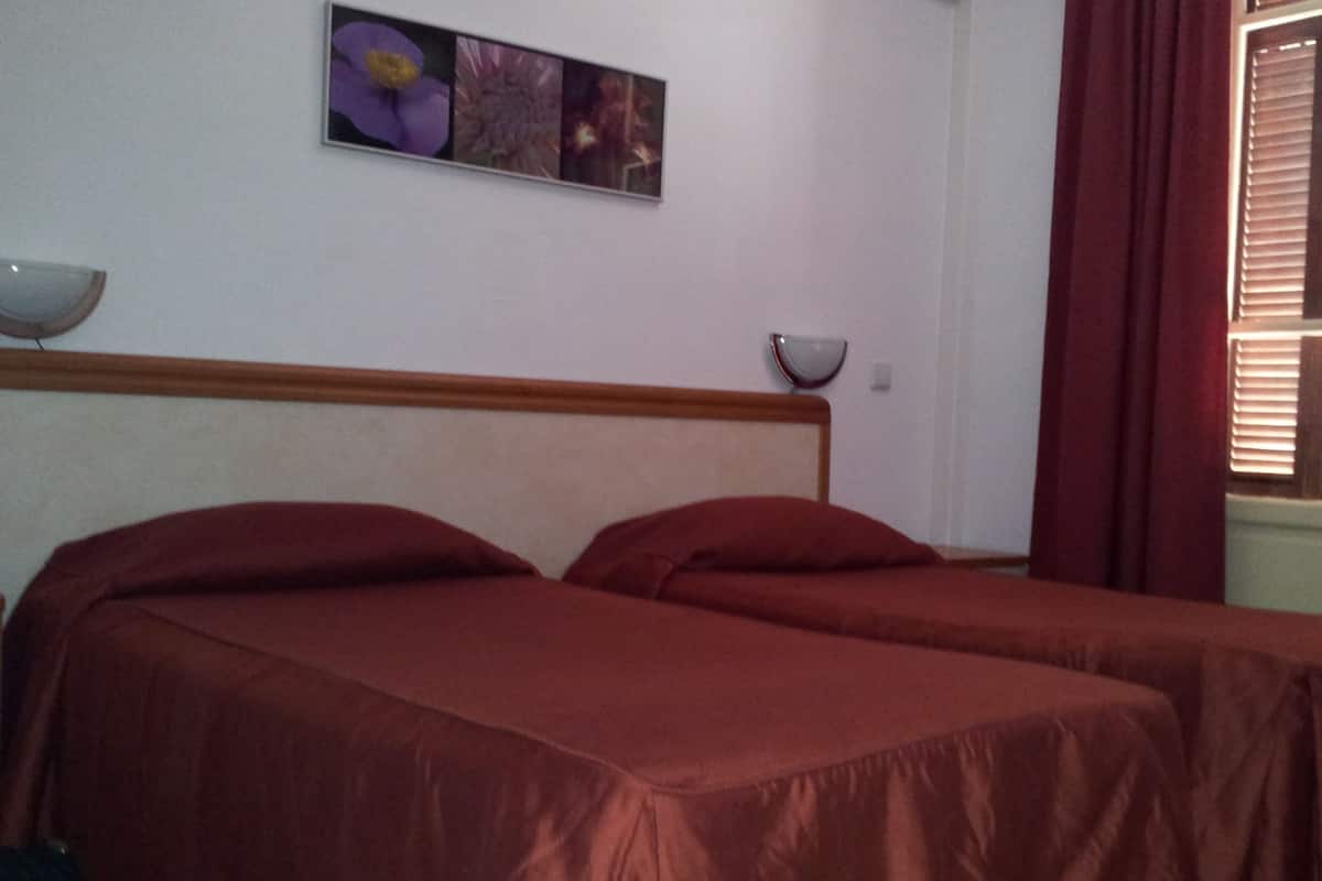 A room with two beds, a wall-mounted picture, a window with blinds, and a bedside table with a lamp.