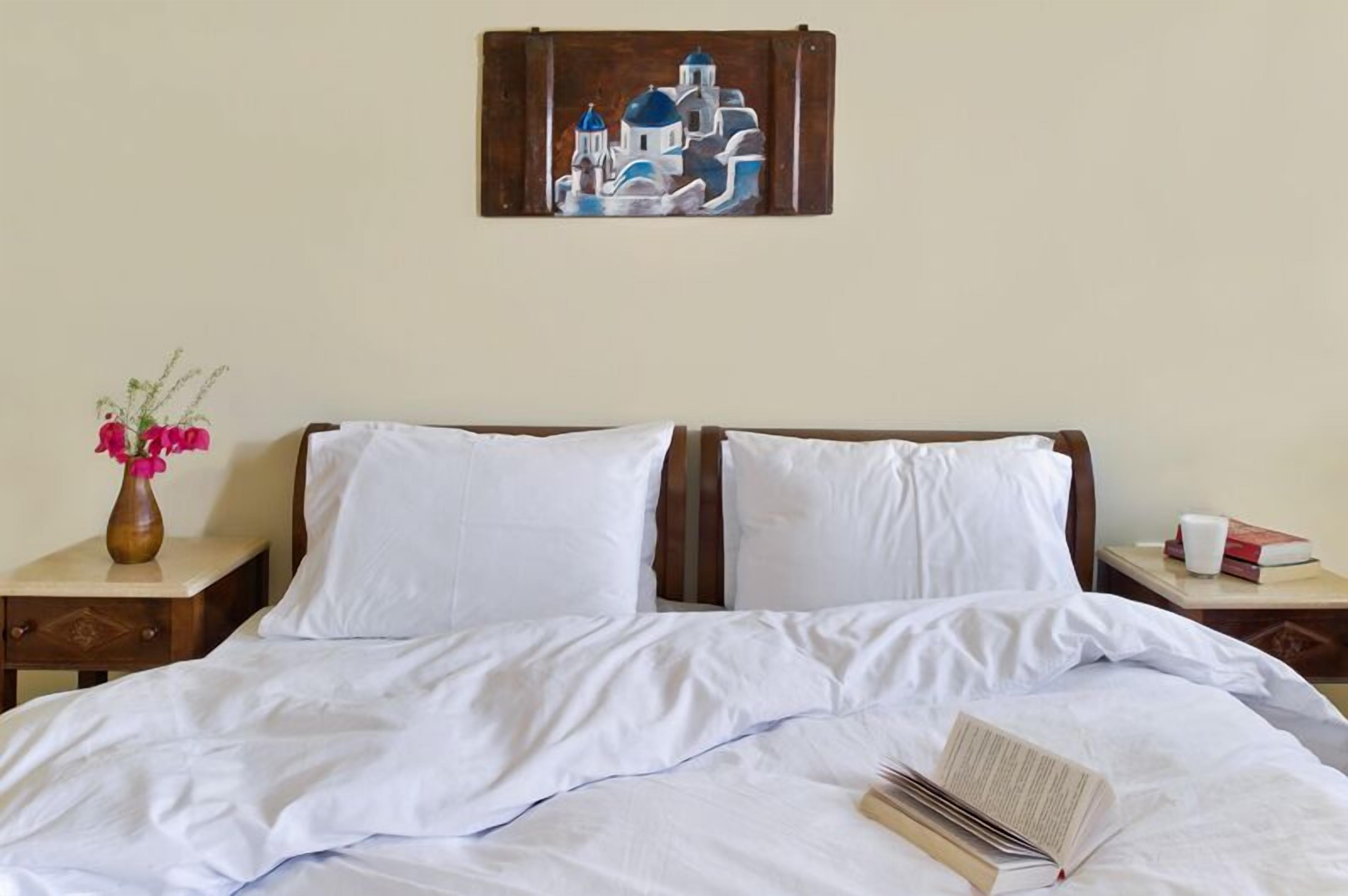 A double bed with white linens, a vase with pink flowers, and a painting of blue and white buildings on the wall.