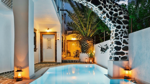 A swimming pool with a stone wall and a palm tree, a door, and a seating area with a lamp. - Galatia Villas (Santorini)