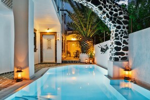 A swimming pool with a stone wall and a palm tree, a door, and a seating area with a lamp. - Galatia Villas (Santorini)