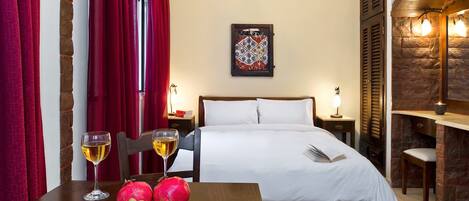 A hotel room with a bed, a table with fruit and wine glasses, a painting on the wall, and a stone fireplace.