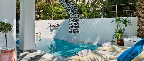 Una piscina infinita con un diseño de arco de piedra único, un área de descanso con un sofá blanco y cojín azul, y una planta en maceta.