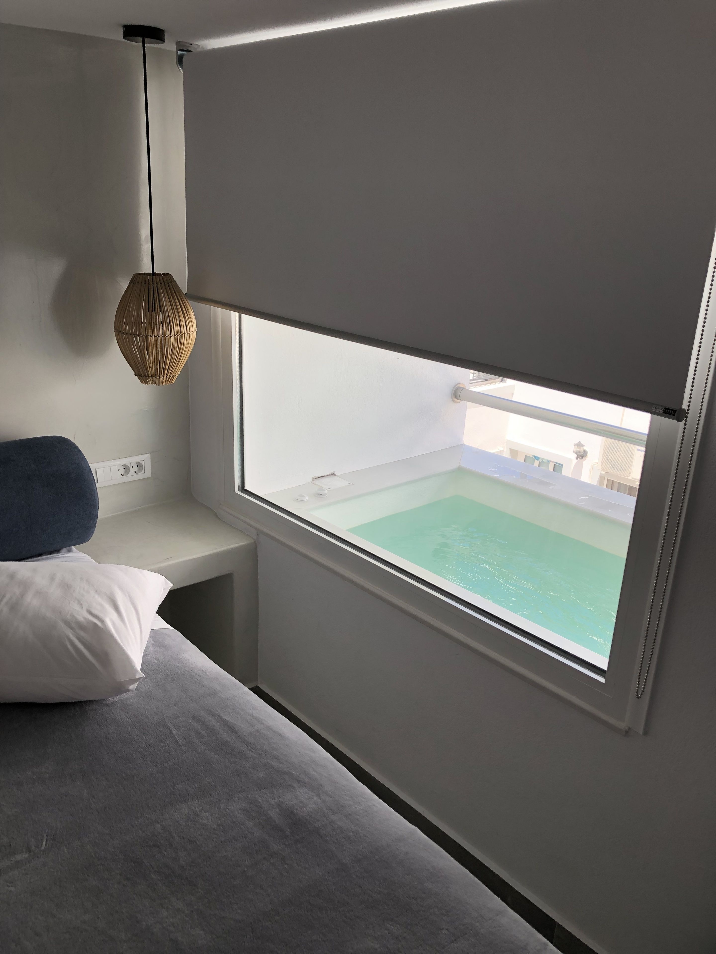 Quarto com uma grande janela, uma cama, uma mesinha de cabeceira e um lustre.