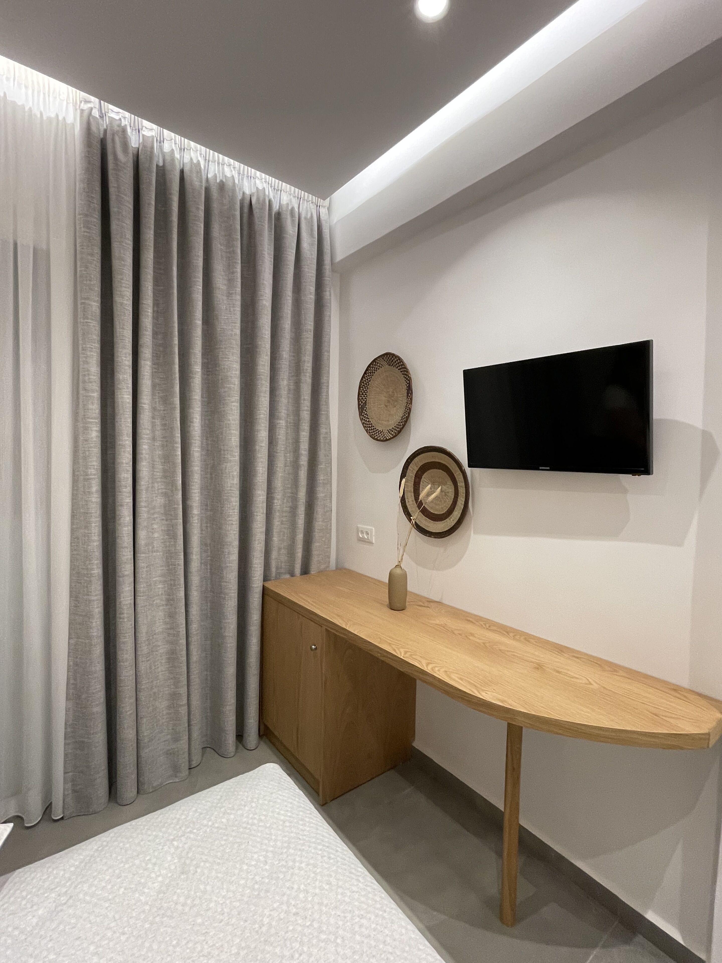 Habitació moderna amb escriptori de fusta, televisió pantalla plana penjada a la paret i finestra gran amb cortines grises.