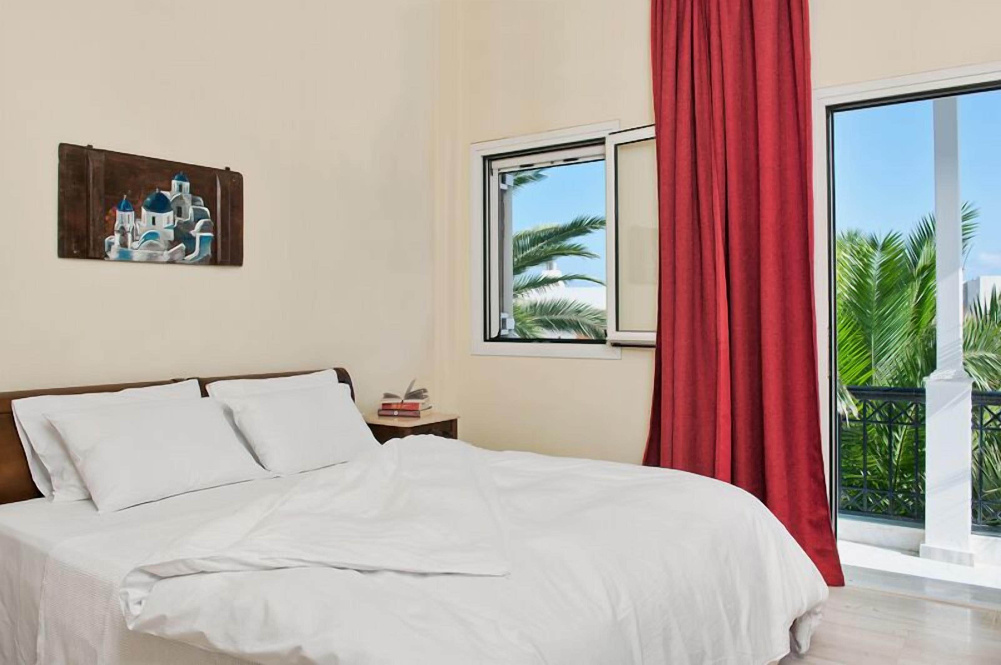 A bedroom with a bed, a nightstand, a painting, a window with a view of palm trees, and a red curtain.