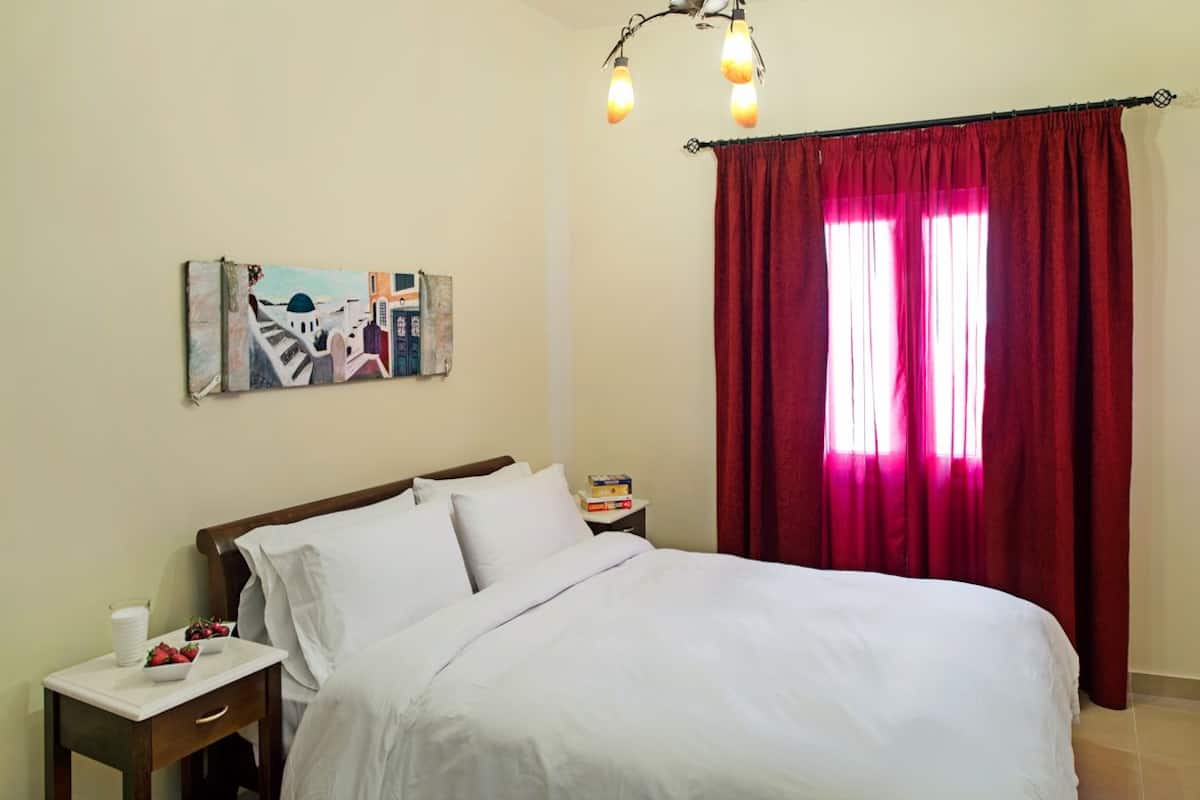 Een net opgemaakt bed met witte lakens, een nachtkastje met een boek en een fruitschaal, een schilderij aan de muur en een raam met rode gordijnen.