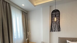 A modern hotel room with a round wooden table, two wicker chairs, a bed with a patterned pillow, and a unique black pendant light. - Galatia Villas (Santorini)